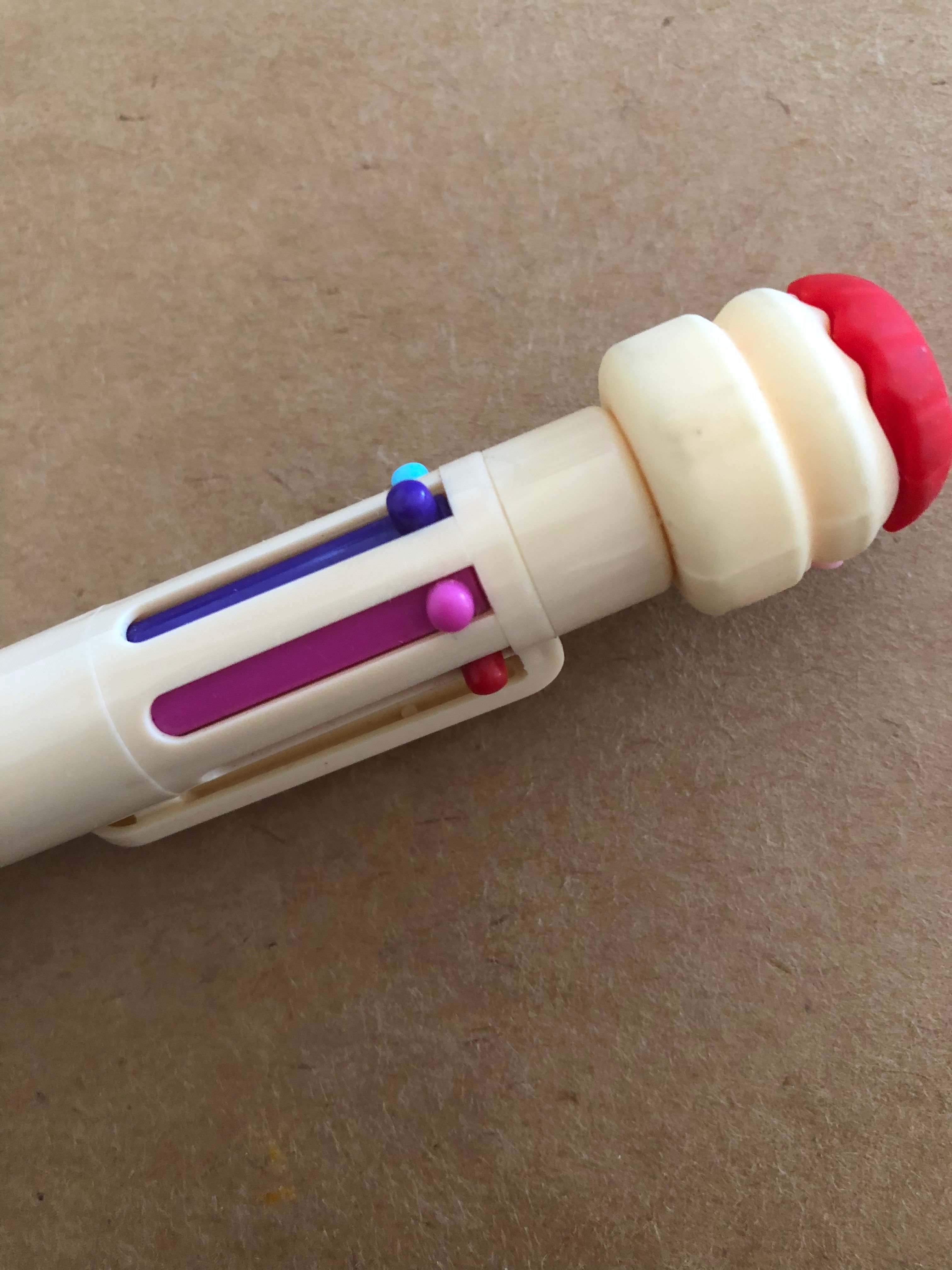 a playful pale yellow pen with a burger shaped top. The burger has a red top and the pen contains multiple writing color choices. Starting from the left there is a light blue, dark purple, magenta pink, and dark red. The pen is lying horizontally, at an angle, on a cardboard surface.