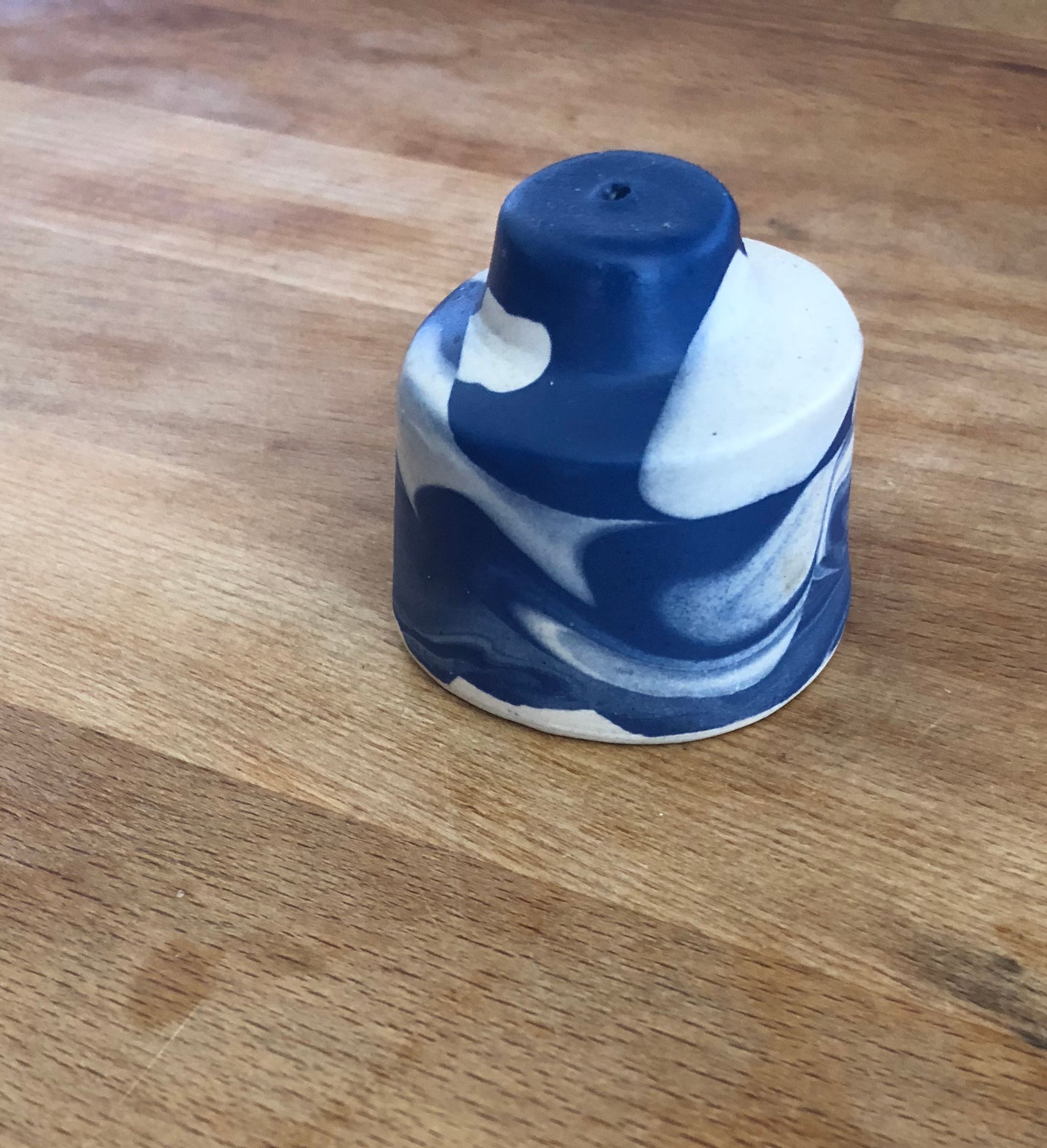 A small ceramic salt shaker made of a swirling dark blue and white gradient. The shape of the salt shaker is not symmetrical and it is sitting on top of a light wooden surface.