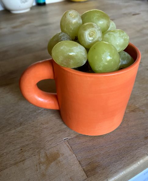 an orange ceramic mug built by the artist Hotel Ceramics sitting on top of an oak wooden counte. The cup is filled to the brim with plump green grapes.