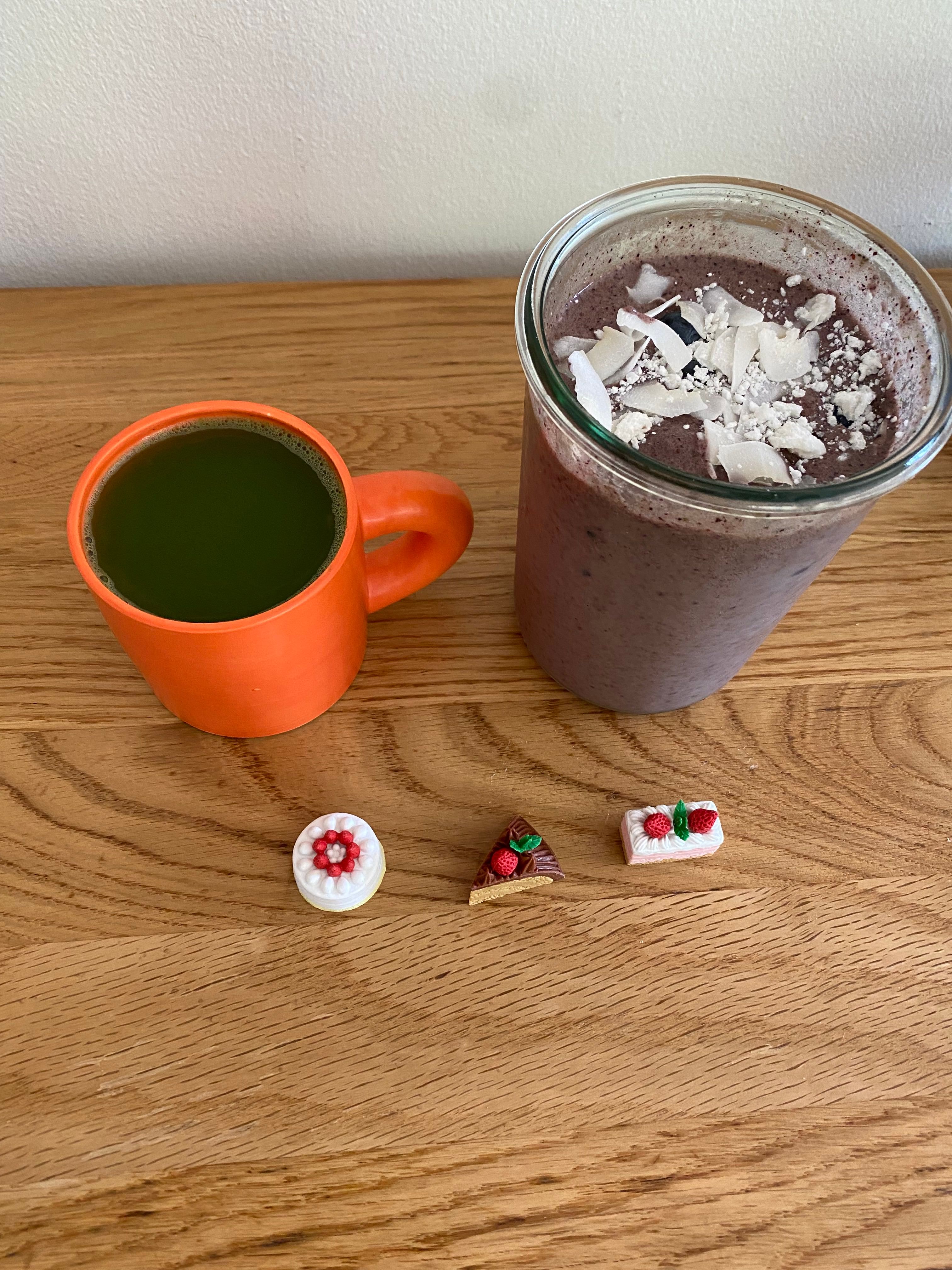 A bright orange ceramic mug with dark green tea is resting on top of a wooden table. To the right of the mug there is a wide rimmed glass jar with a purple smoothie inside of it topped with coconut shreds. In front of the two beverages there are three small erasers each shaped like miniature pieces of cake.