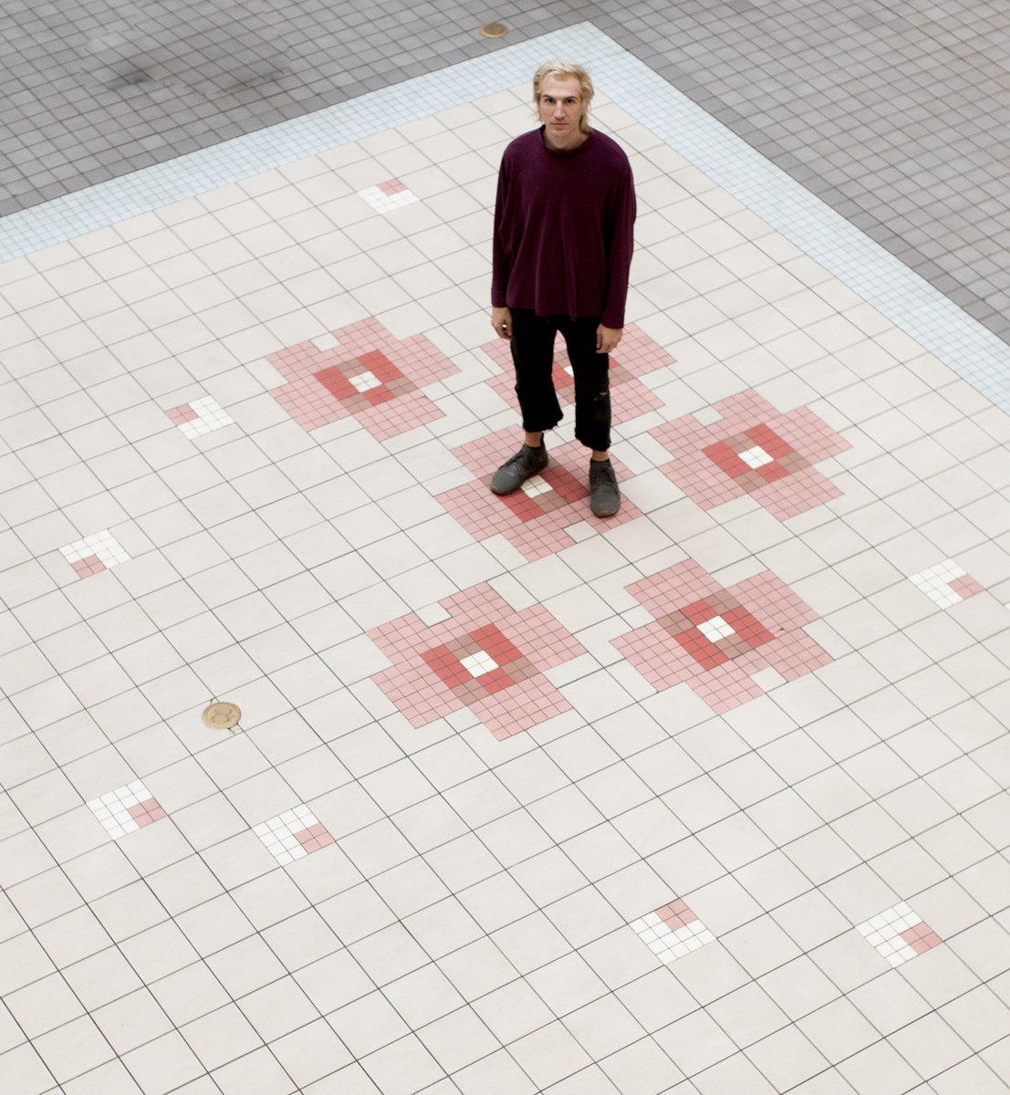 A bird's eye view of Brin, standing in the center of a tiled floor. The floor has small pink, terra cotta, and bright white tiles creating square like shapes and designs. Brin has blonde platinum hair and is wearing a burgundy velour long sleeve top, black pants, and black shoes. He is looking up at the camera