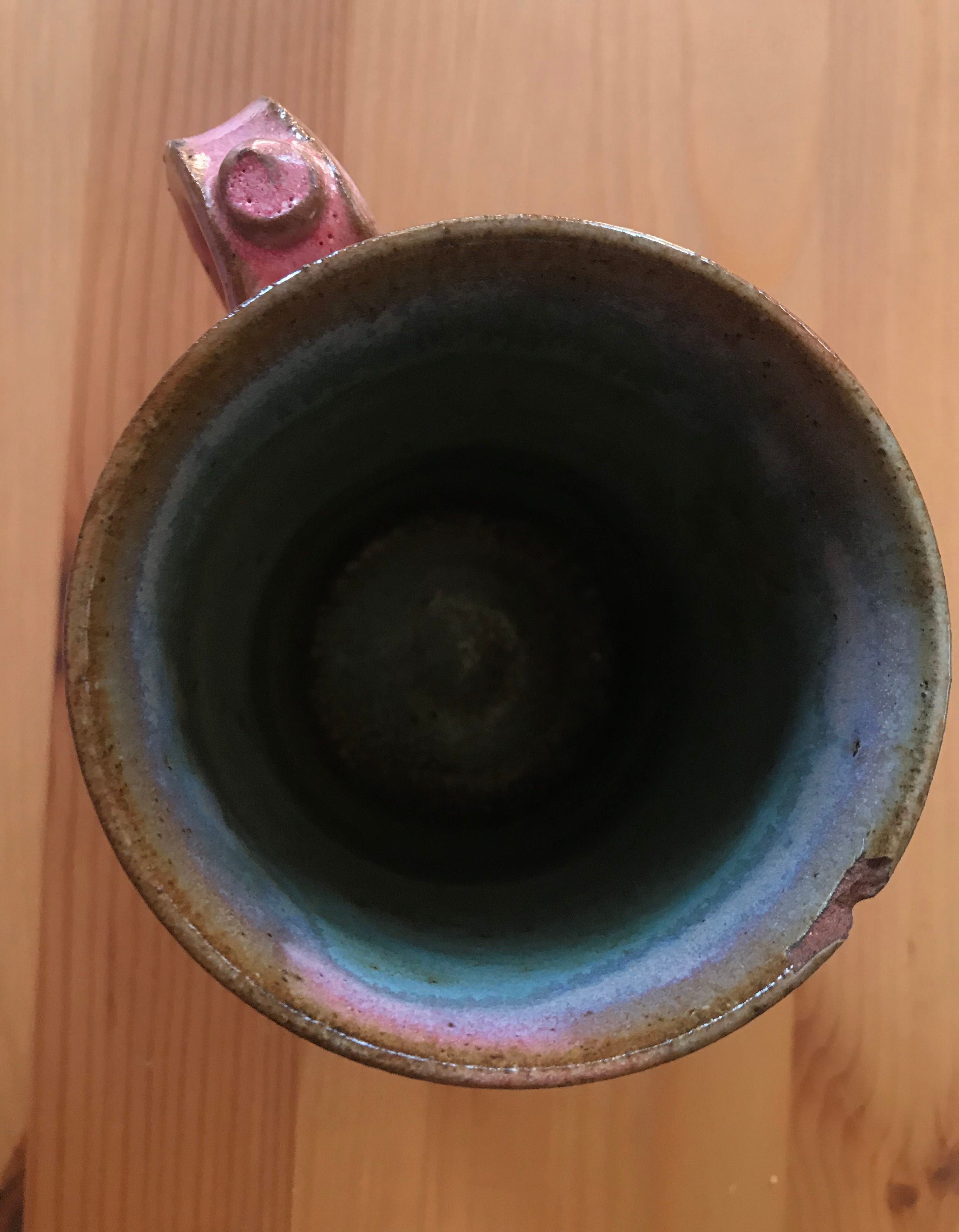 A close up bird's eye view of Ghorba's ceramic pink and blue mug sitting on top of a maple wood surface. The inside of the mug is dark blue with slight ridges going all the way through the inside of the mug. There is a small chip on the right side of the rim of the mug and it is has a brown glaze. The handle of the mug is bubblegum pink and has a small swirl shape on it.