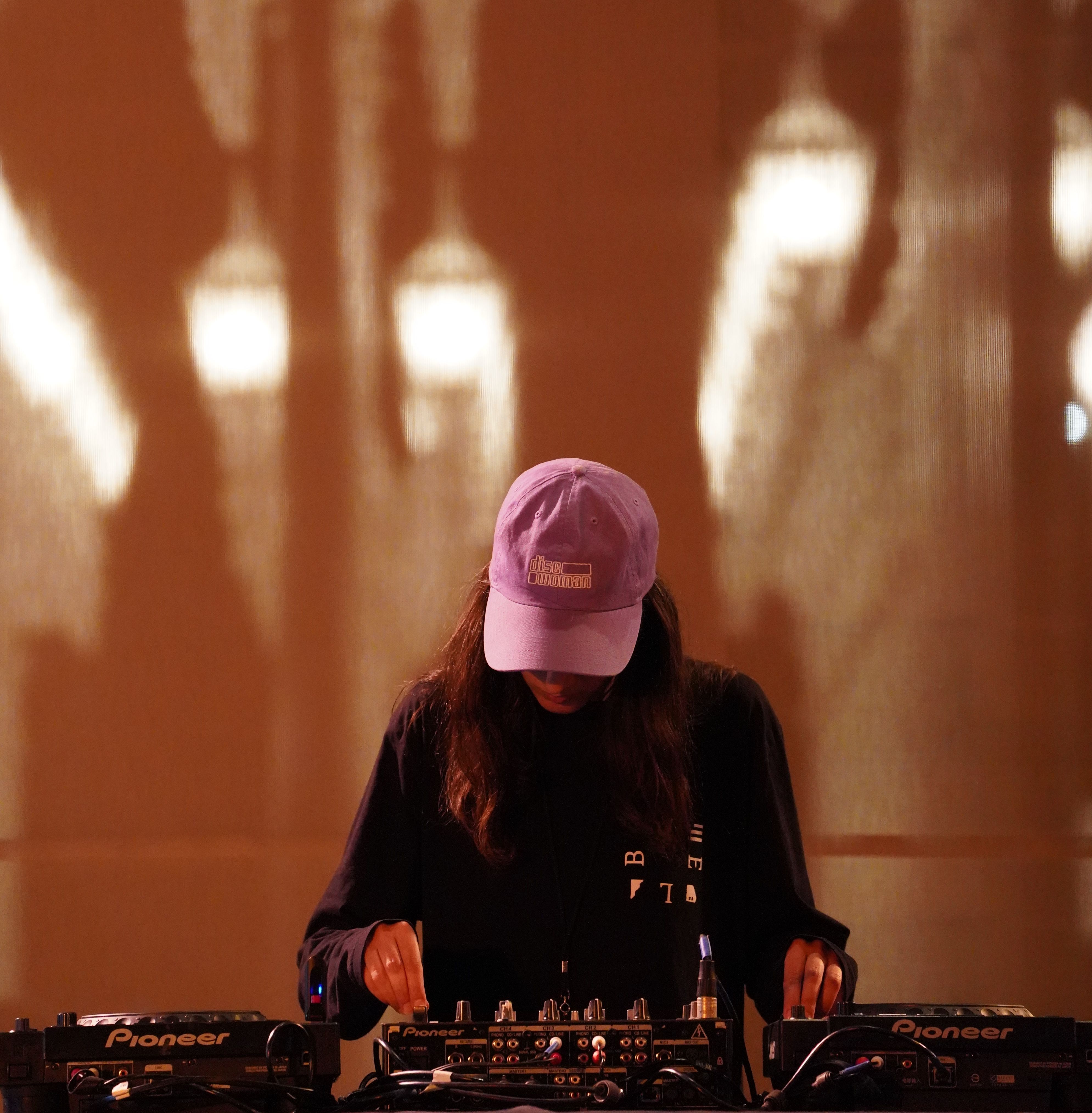 Maral is standing behind a CDJ DJ setup. She is looking down at the DJ mixer, adjusting knobs with her right hand while the other is placed on the CDJ. She has long brown/black hair, a long sleeve black top on and a light pink hat on her head. Her background is a blurry image of the lights in front of LACMA: many white tall street lights in a row.