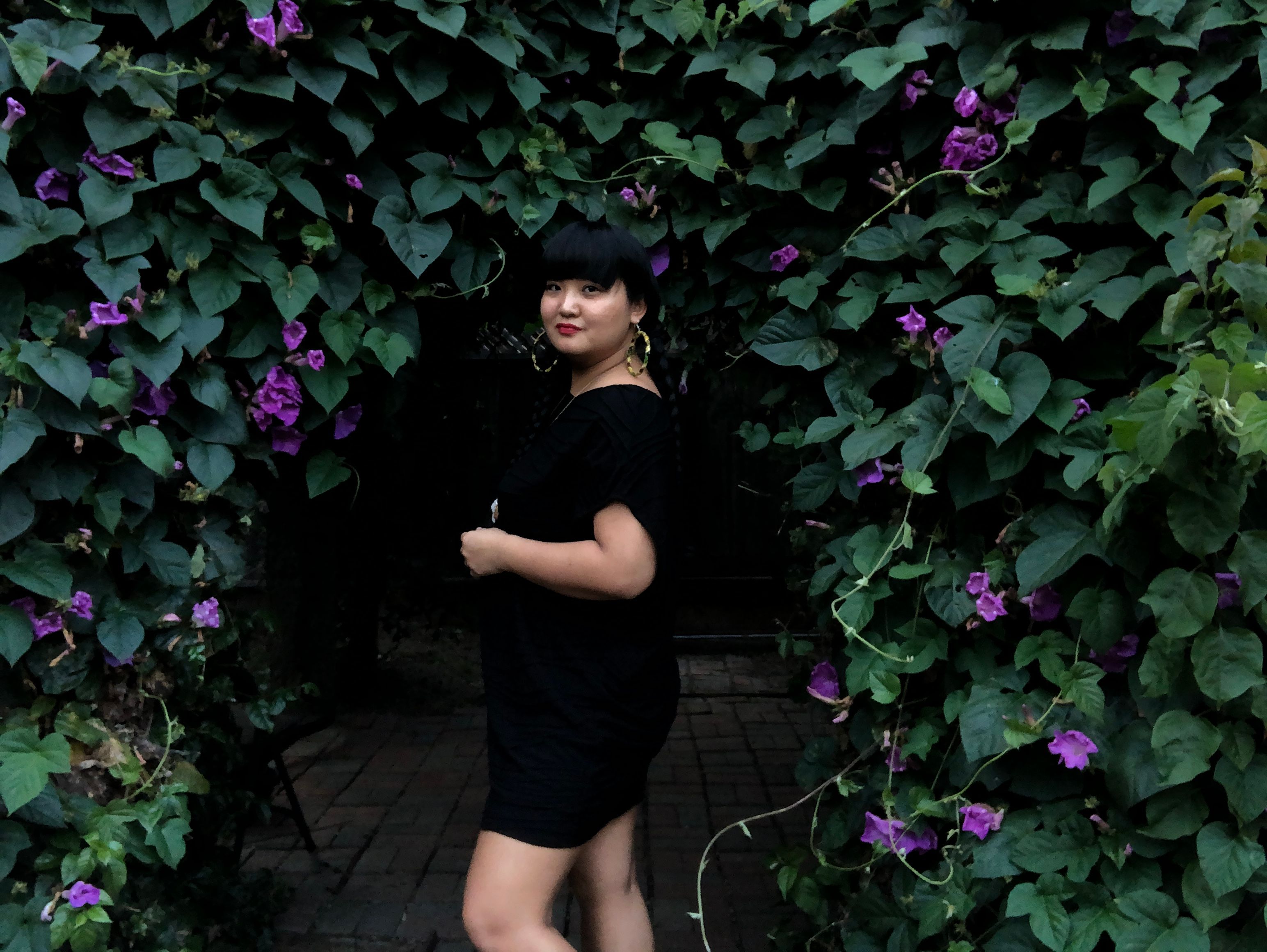 T-Kay wearing a short sleeved black dress, large hoops, and red lipstick turned to the side and standing against a backdrop of morning glory vines