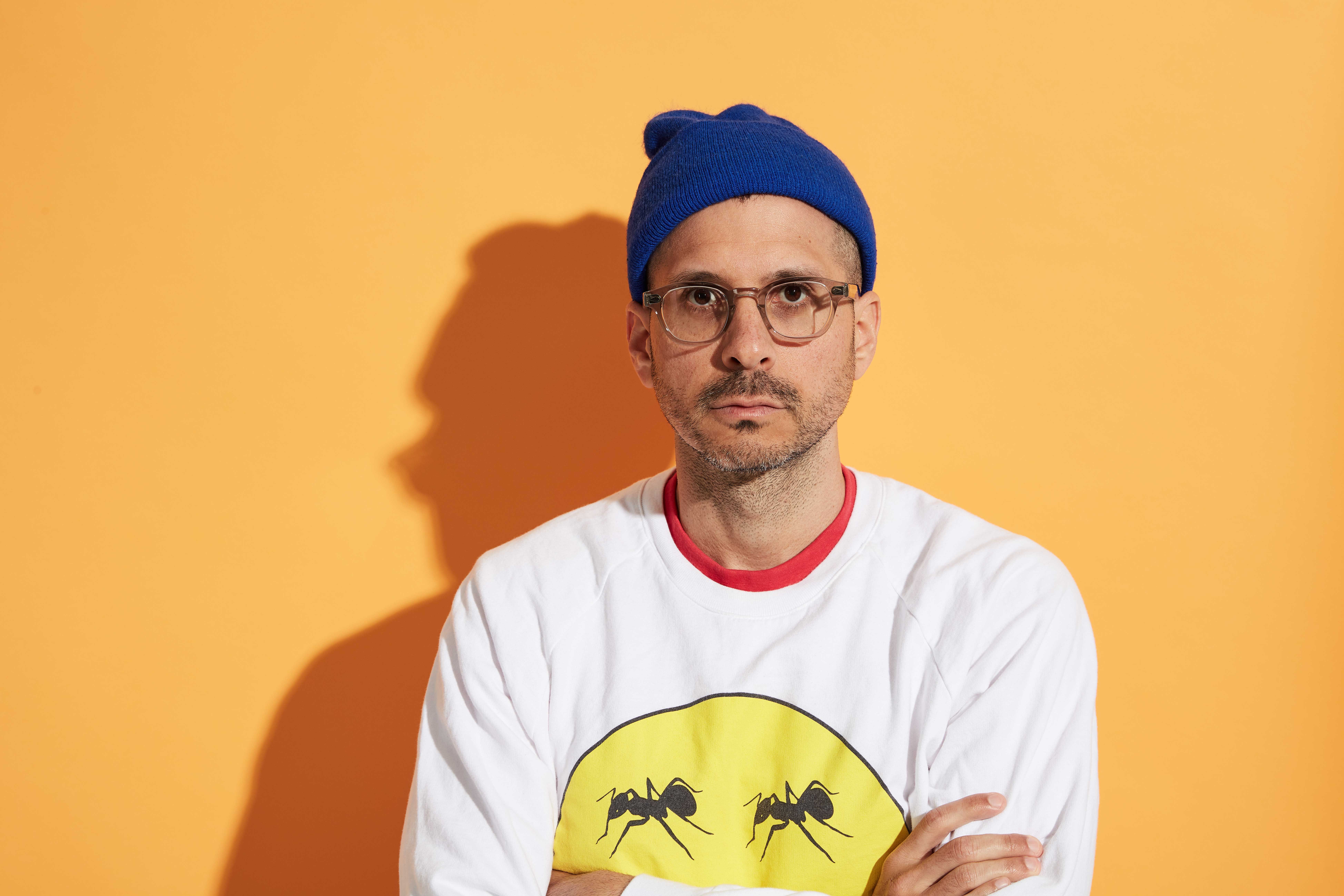 Yoni Wolf is in front of a bright yellow background. He is wearing a white sweatshirt with a yellow circle on it with two ants within the circle. He has a red shirt showing from underneath his sweatshirt and is wearing glasses with a translucent brown rim and a bright blue beanie.