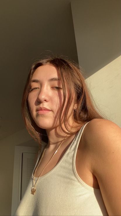 Kelsey, a light skinned person is looking down at her camera and her face is illuminated by natural lighting. She is wearing a white ribbed tank top and a gold necklace with a gold charm which reads "sober brat". She is situated in front of an off white/cream wall and white door.