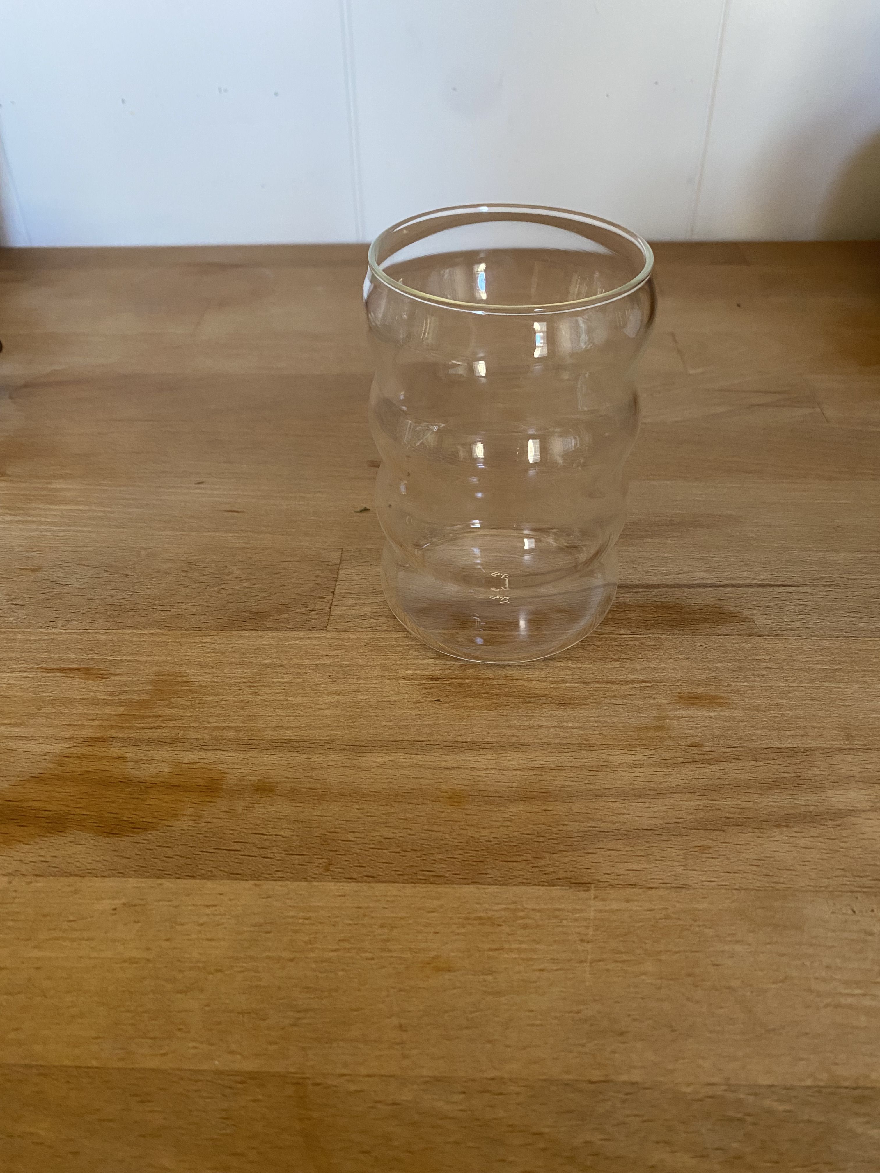 A glass cup sitting on top of a light wooden surface. The cup has ripples in it and is wavy in nature. 