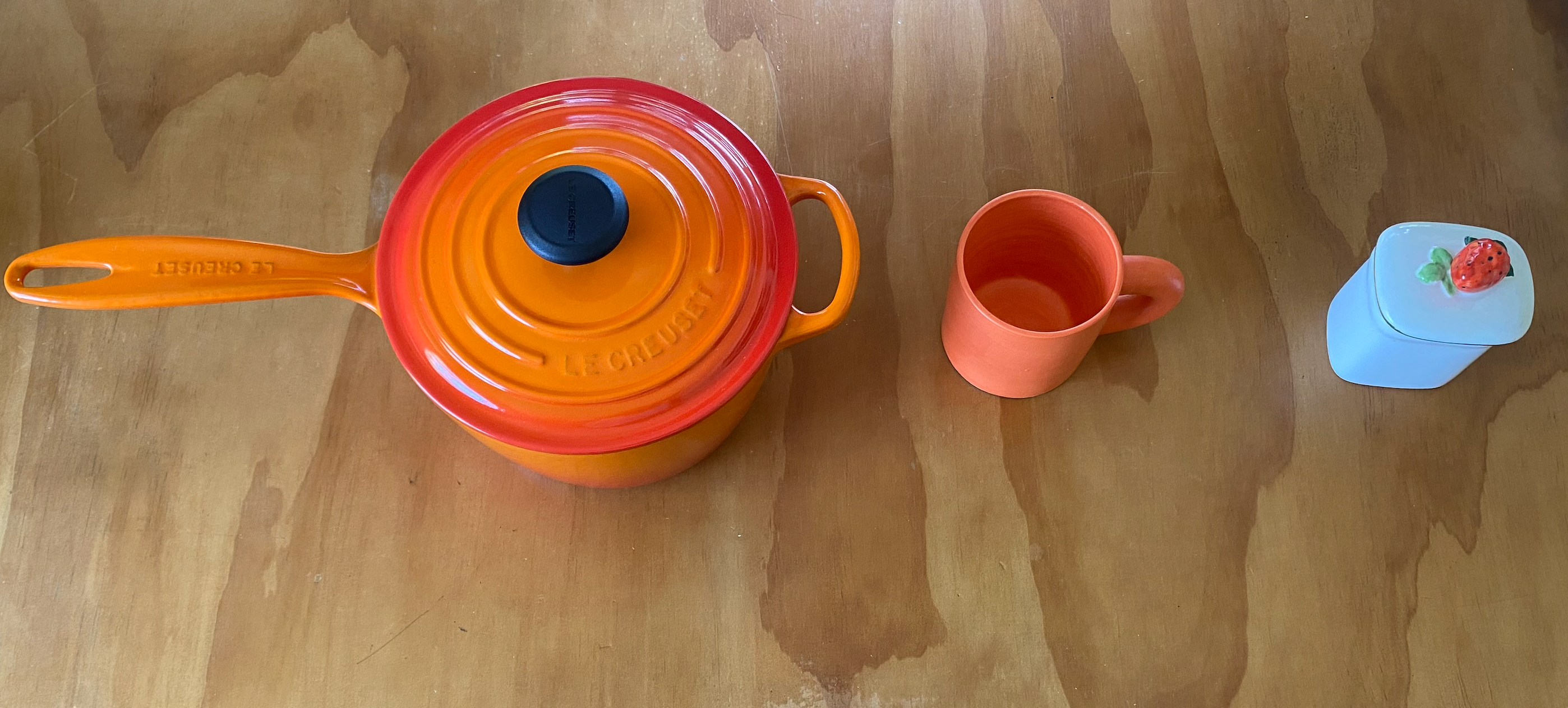 Three vessels on top of a light wood surface. From left to right there is a Le Creuset medium bright orange sauce pan with a red to orange inward moving gradient on the lid, with a black handle. Then there is a small orange ceramic mug with a thick half circle handle. To the right of the mug is a small porcelain jelly container with a ceramic strawberry on the lid.