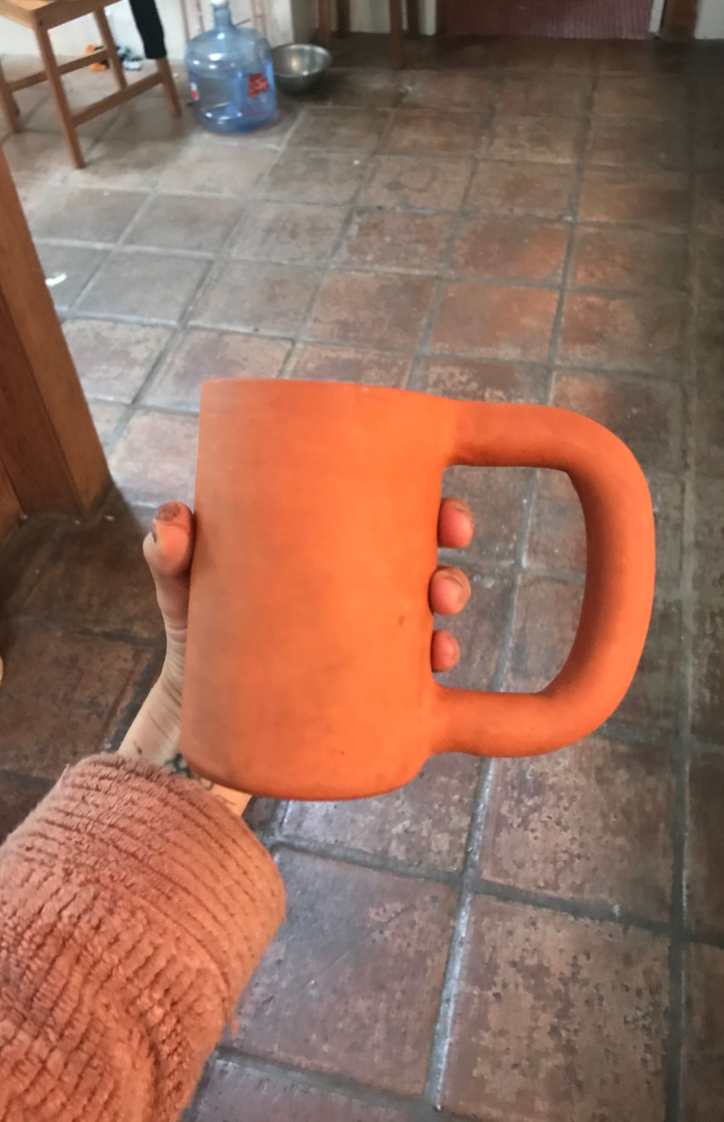 Fino is outstretching their hand and is holding a large ceramic terra cotta mug which has a large oversized handle on it's right side. Fino has a light brown sleeve. In the backgeound of the photo there are light brown / grey tiles and a wooden chair and large water jug visible in the background. 