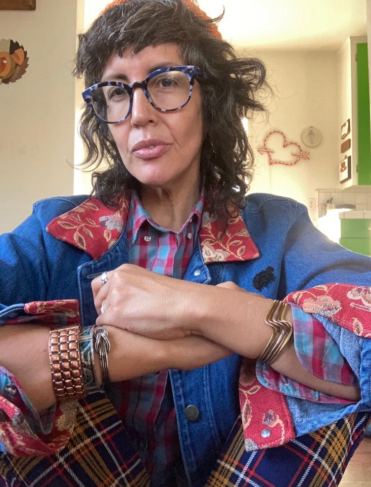 Vanessa is sitting in front of the camera, head slightly angled upward and to the side. They are wearing a vibrant jean button up long sleeve shirt with a red collar and floral print cuffs. The jean top is over a blue and magenta flannel top. Vanessa's arms are crossed, showing their gold, copper, and silver bracelets. Their pants are blue, red, white, and yellow flannel print and their glasses are large with a dark gray and white print. Vanessa's hair is wavey and to their shoulders with an orange knit stalking cap on their head. In the background you can see pale yellow walls and light green cupboards. There is a heart and arrow decoration hung on the wall.