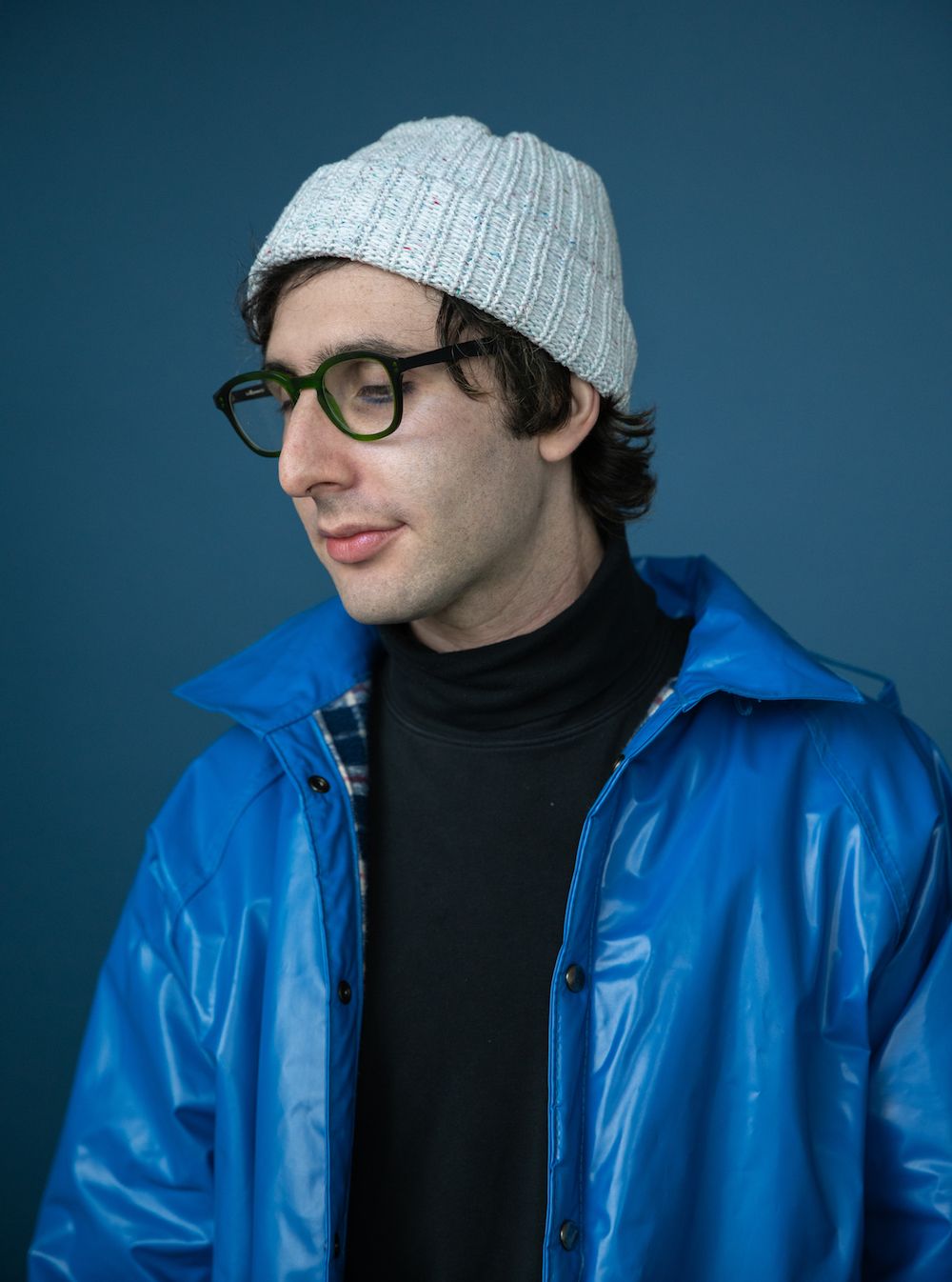 person against a blue backdrop with light skin, dark hair, green glasses, a white beanie, a black turtleneck, and a blue jacket.