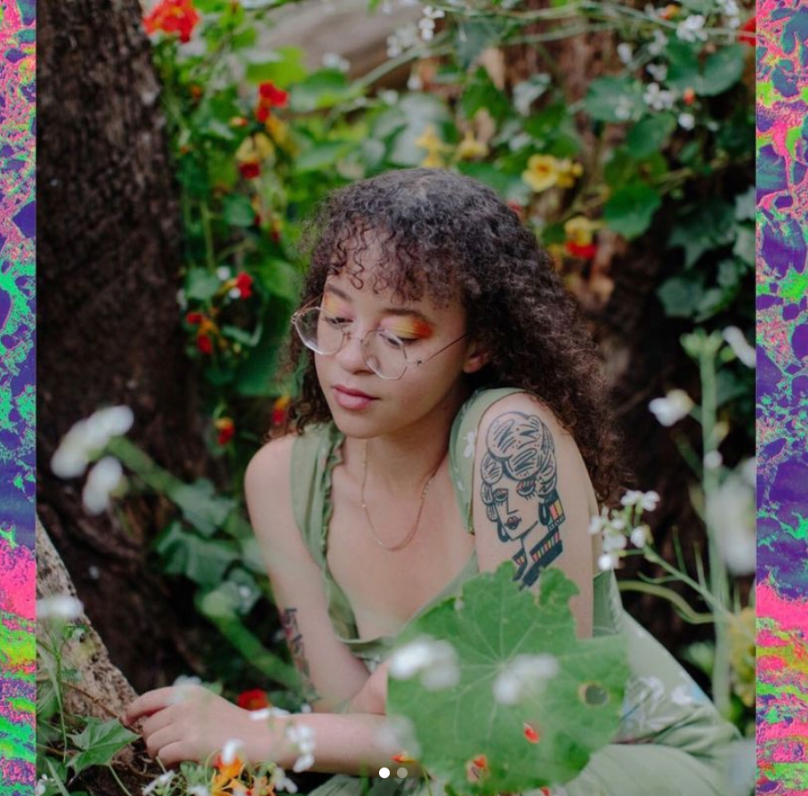 A photo of Xyla in which they are in a garden of vibrant green, yellow, white, and, orange flowers and leaves. They are looking wearing a light green clothing article with tank top sleeves and a gold delicate gold necklace. They have a tattoo on their left arm of a person's head and neck. Xyla has circular clear glasses on and is wearing bright orange eye shadow. The photo is overlaid onto a psychedelic background made up of neon purples, greens, and pinks.   