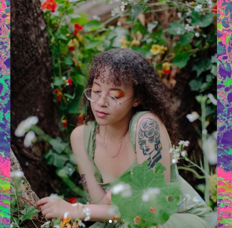A photo of Xyla in which they are in a garden of vibrant green, yellow, white, and, orange flowers and leaves. They are looking wearing a light green clothing article with tank top sleeves and a gold delicate gold necklace. They have a tattoo on their left arm of a person's head and neck. Xyla has circular clear glasses on and is wearing bright orange eye shadow. The photo is overlaid onto a psychedelic background made up of neon purples, greens, and pinks.