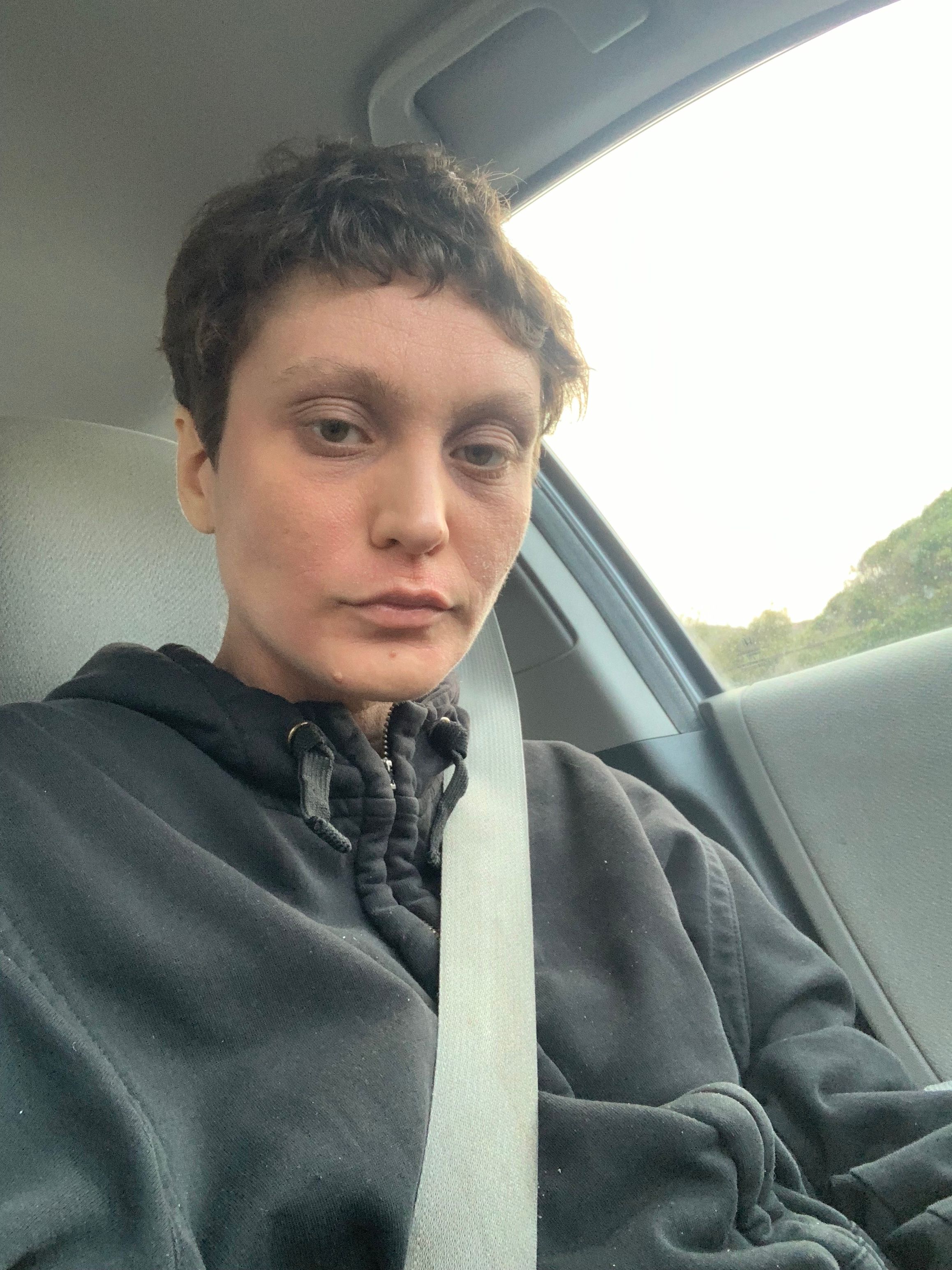 A selfie of a light skinned person with short dark brown hair. They are wearing a black hooded jacket that is zipped up. They are sitting in the grey interior of a car with a seatbelt across their chest. There is a lot of light coming from the car window behind them.