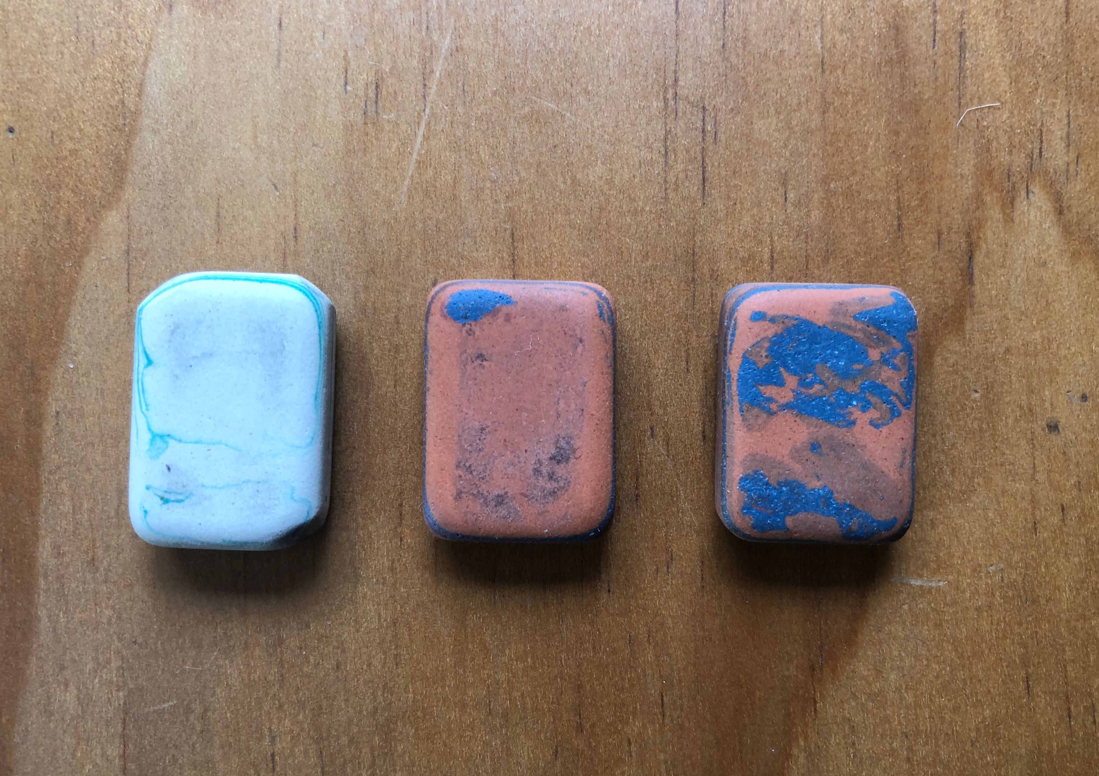 three small rectangle shaped erasers resting on top of a light wood counter top. From viewer's left to right the erasers are white with blue marbling, terra cotta with blue marbling and the third one is also terra cotta with blue marbling.