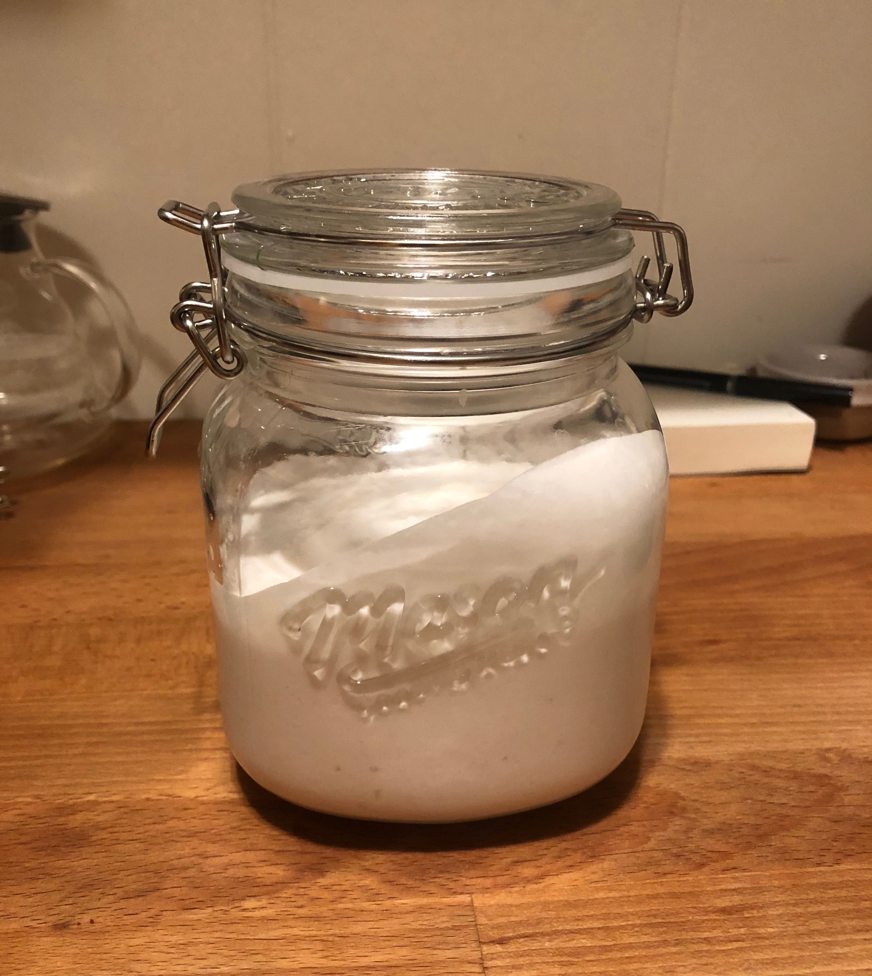 A medium size glass jar with a metal clasp top which is closed. Inside the jar there is white creamy yogurt filling about three fourths of the jar. On the front of the jar it reads "Mason" indented into the glass. The jar is sitting on top of a walnut wood counter.