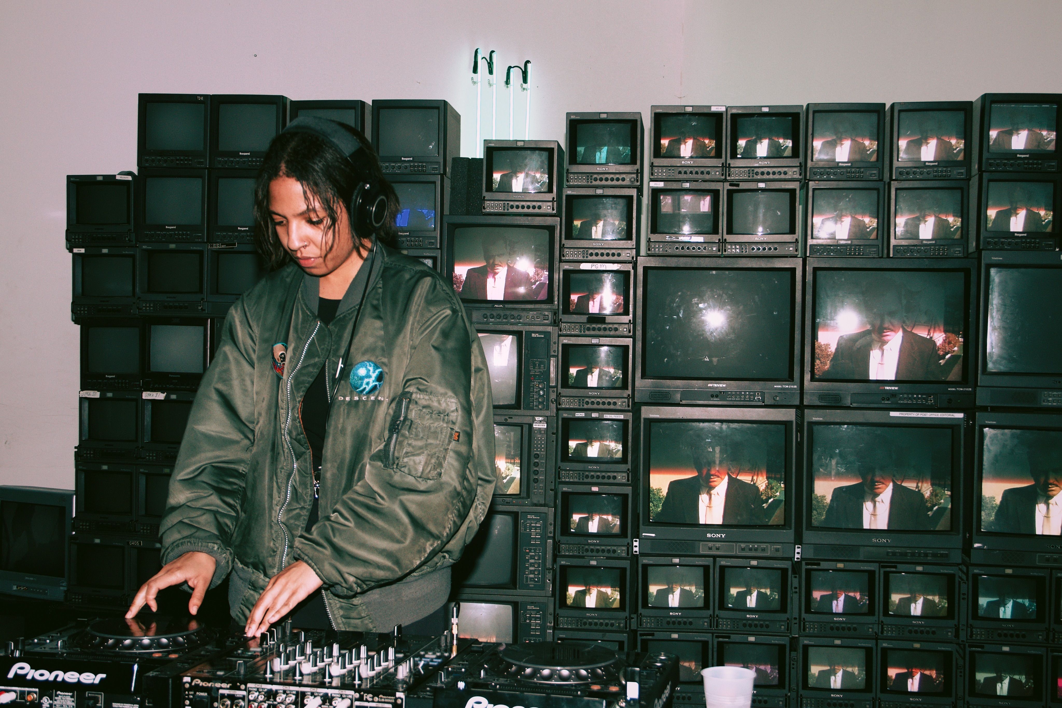 Bapari djing with CDJs standing in front of a wall of stacked vintage tv screens. The screens are each playing an image of a man in a tux.