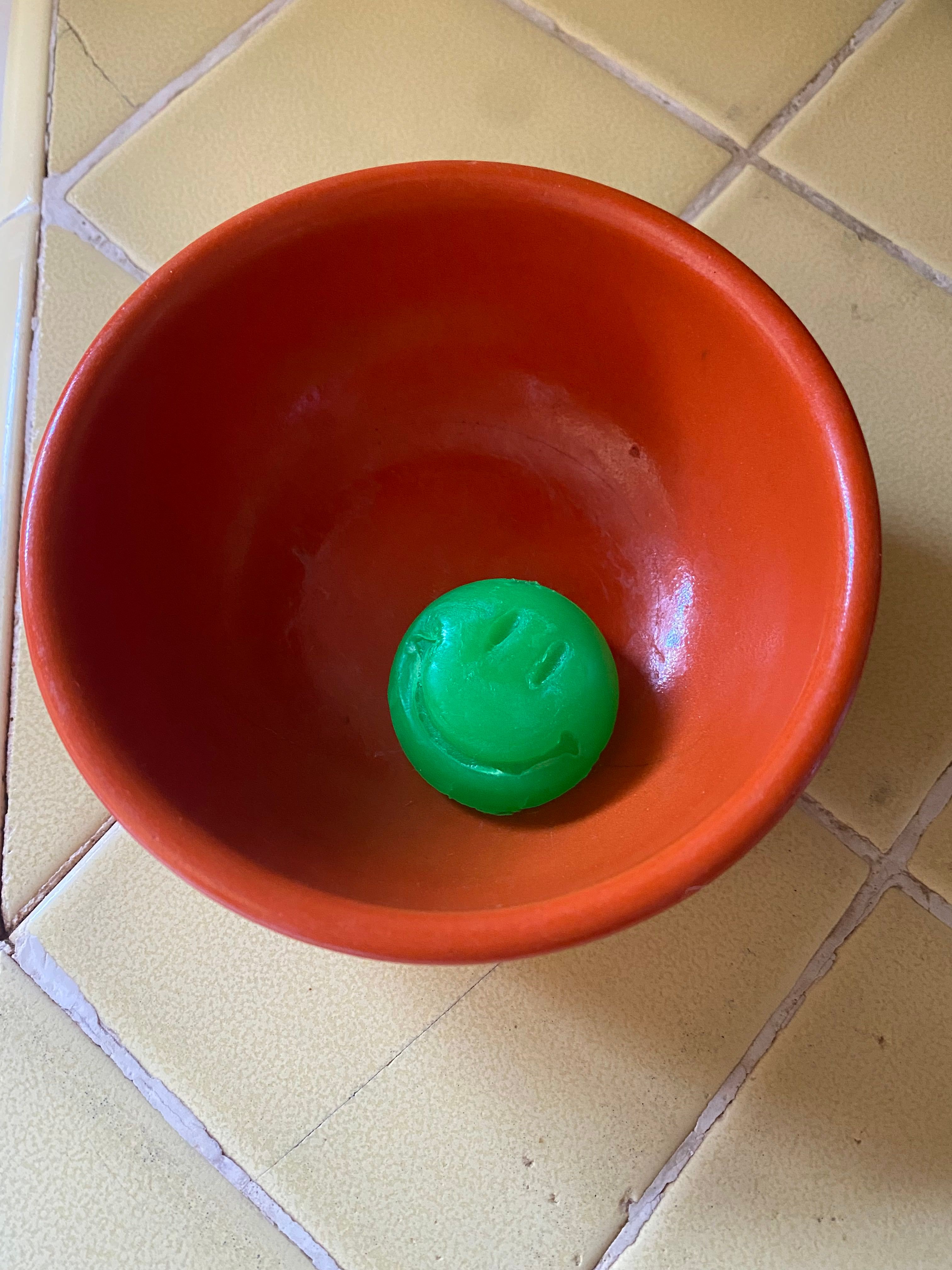 An orange ceramic bowl with a glossy finish sitting on top of a pastel yellow tile countertop. Inside of the green bowl there is a small green soap shaped like a smiley face.