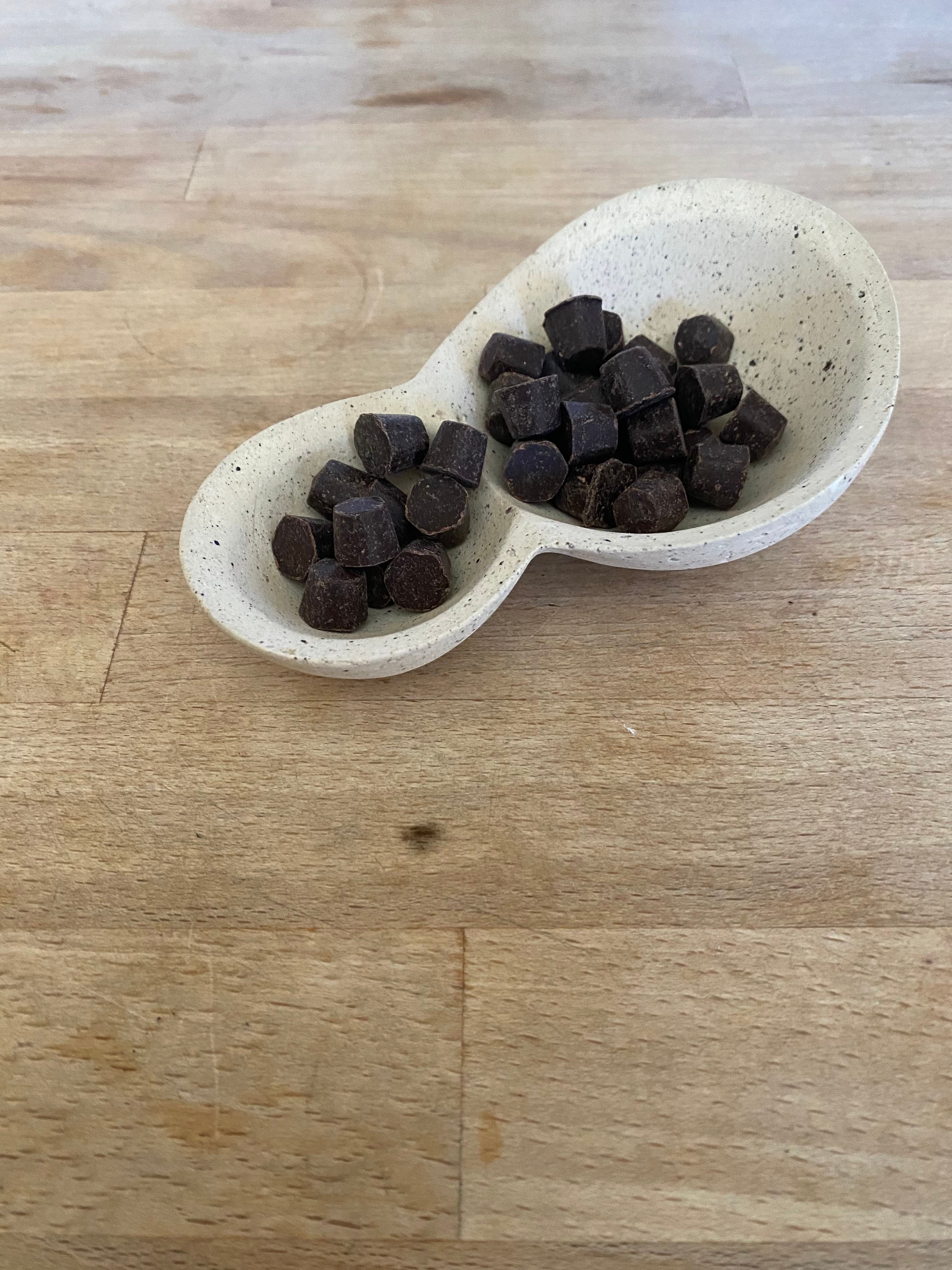 an off white ceramic vessel with brown speckles made of two connected spherical shapes filled with dark chocolate gems. 