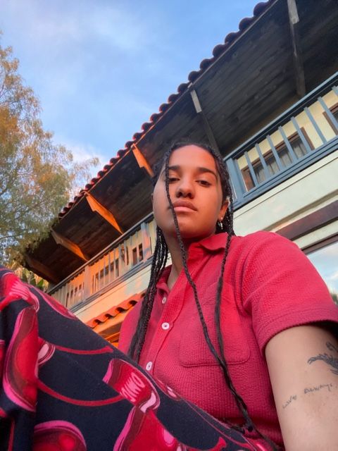 Ella sits outdoors wearing an orange button up with printed red pants while looking down at the camera. She is in front of an off white house with light blue rails. The lighting appears as if the photo was taken during twilight hour.