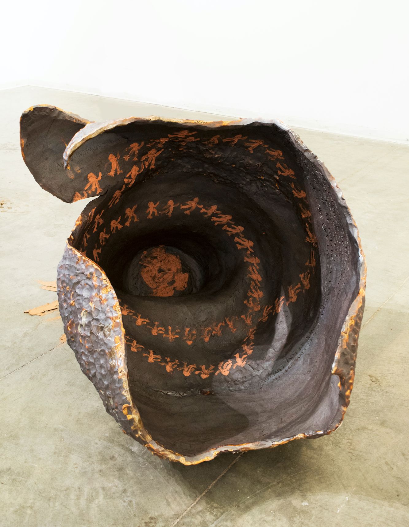 A brown open ceramic cocoon with small orange figures encircling the interior of the vessel. The opening of the vessel looks as though you are peering into the cocoon.