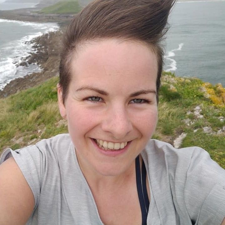 A smiling person taking a selfie on a grassy coastal cliff overlooking the sea. Strong wind lifts their short hair straight upward, creating a playful look. Rocky shoreline, rolling waves, and a cloudy sky form the scenic background.
