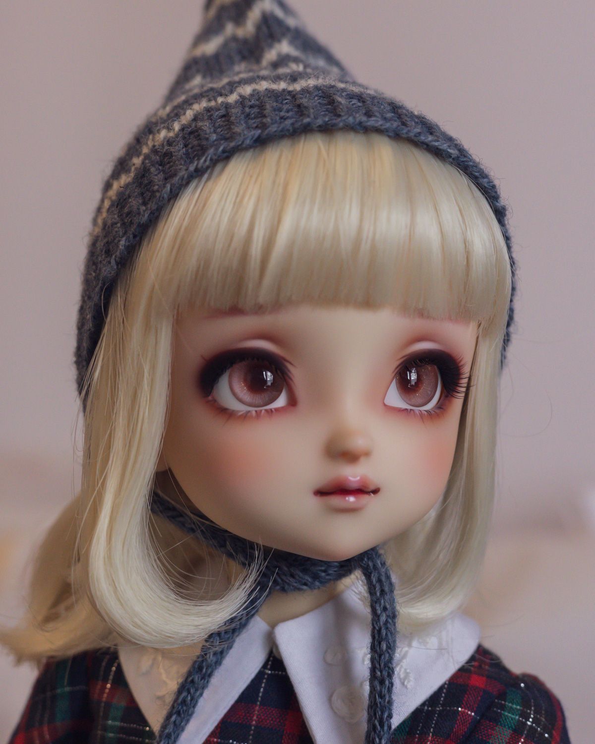 Three-quarter portrait photo of a female ball-jointed doll with big peachy-brown eyes and a blond bob with blunt-cut fringe, wearing a knitted pointed pixie hat that covers her ears. The hat is dark blue with white zig-zag stripes and ties under her chin..