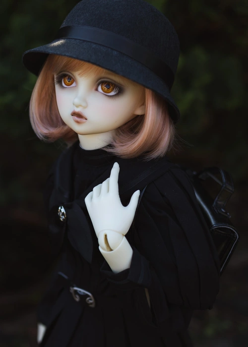 Portrait photo of a ball-jointed resin doll. She is outside in a garden with dark foliage, wearing a black old-fashioned school uniform and a cloche hat. She carries a black school bag on her back. Her resin is pale white, her eyes are orange and her bobbed hair is peach.