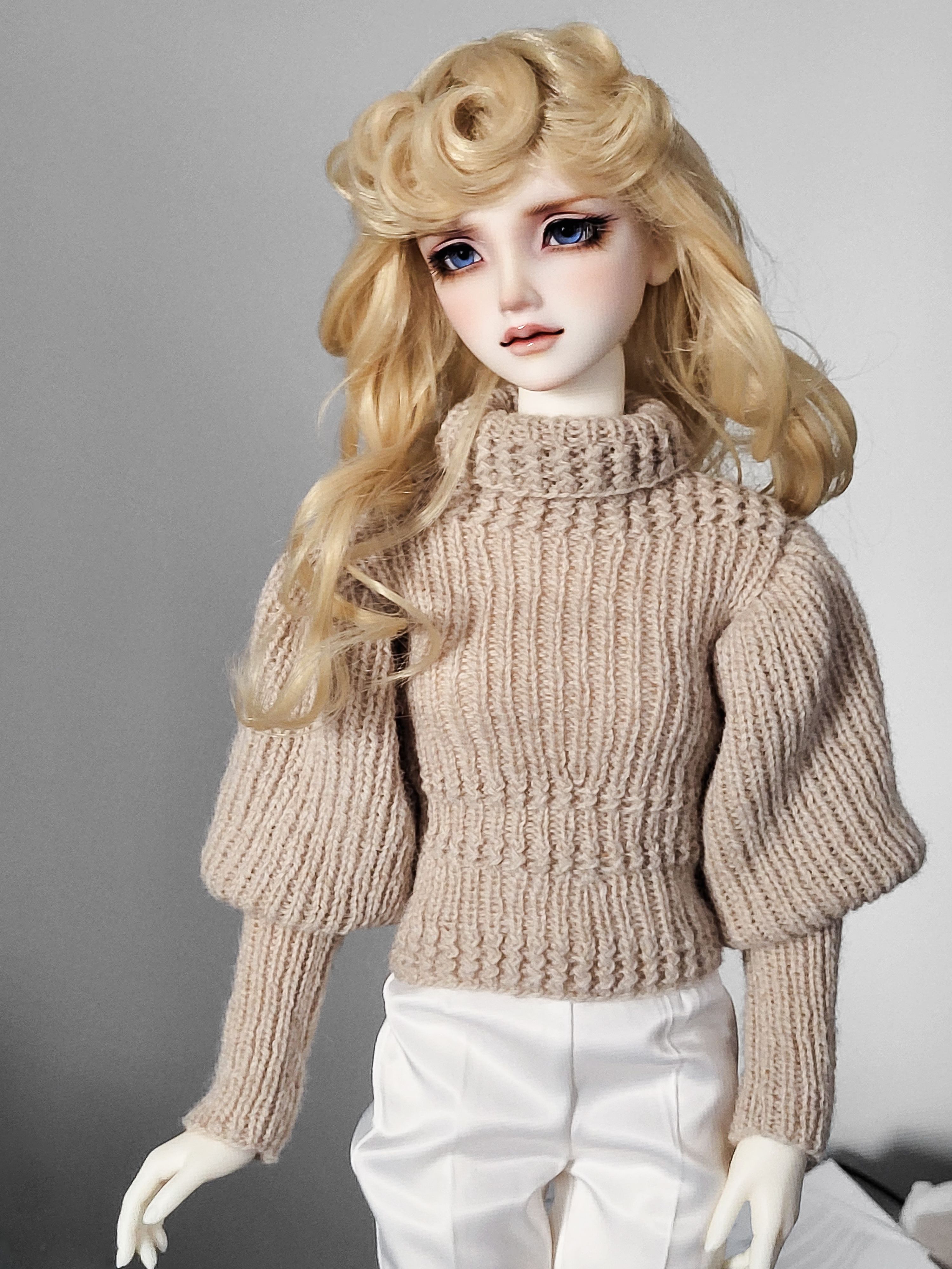 Female resin ball-jointed doll with long curly blond hair, a stern expression, blue eyes, wearing a knitted jumper.