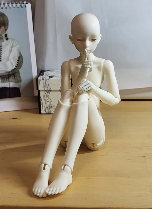 Male ball-jointed doll sitting on a table with knees pulled up, holding a finger to its lips.