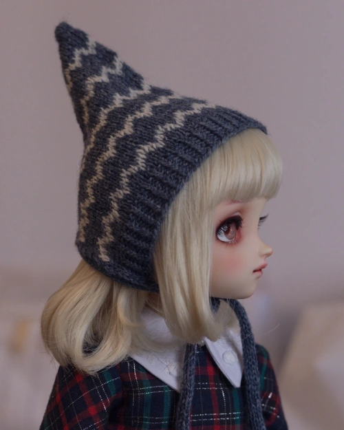 Side-profile photo of a female ball-jointed doll with a blond bob with blunt-cut fringe, wearing a knitted pointed pixie hat that covers her ears. The hat is dark blue with white zig-zag stripes and ties under her chin..