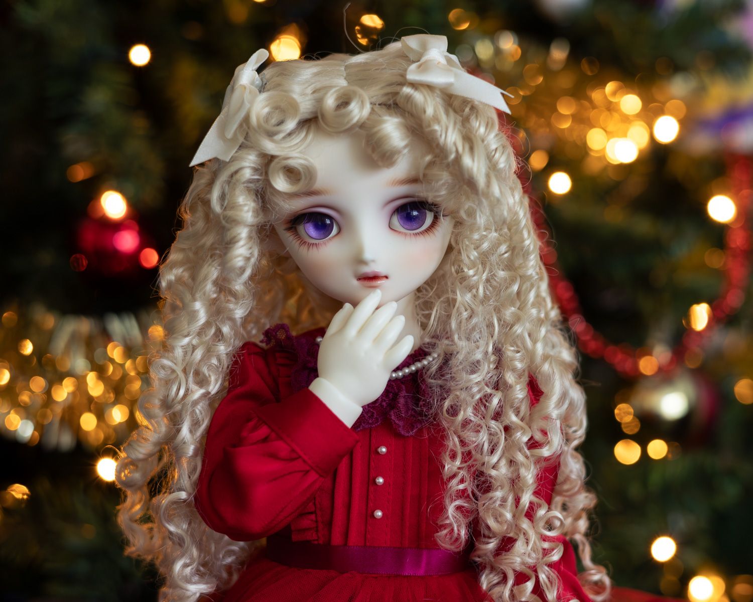 Half-length portrait of a female ball-jointed doll with long blonde hair in ringlets and big purple eyes, wearing a red dress. She has one hand in front of her mouth and behind her is a Christmas tree.