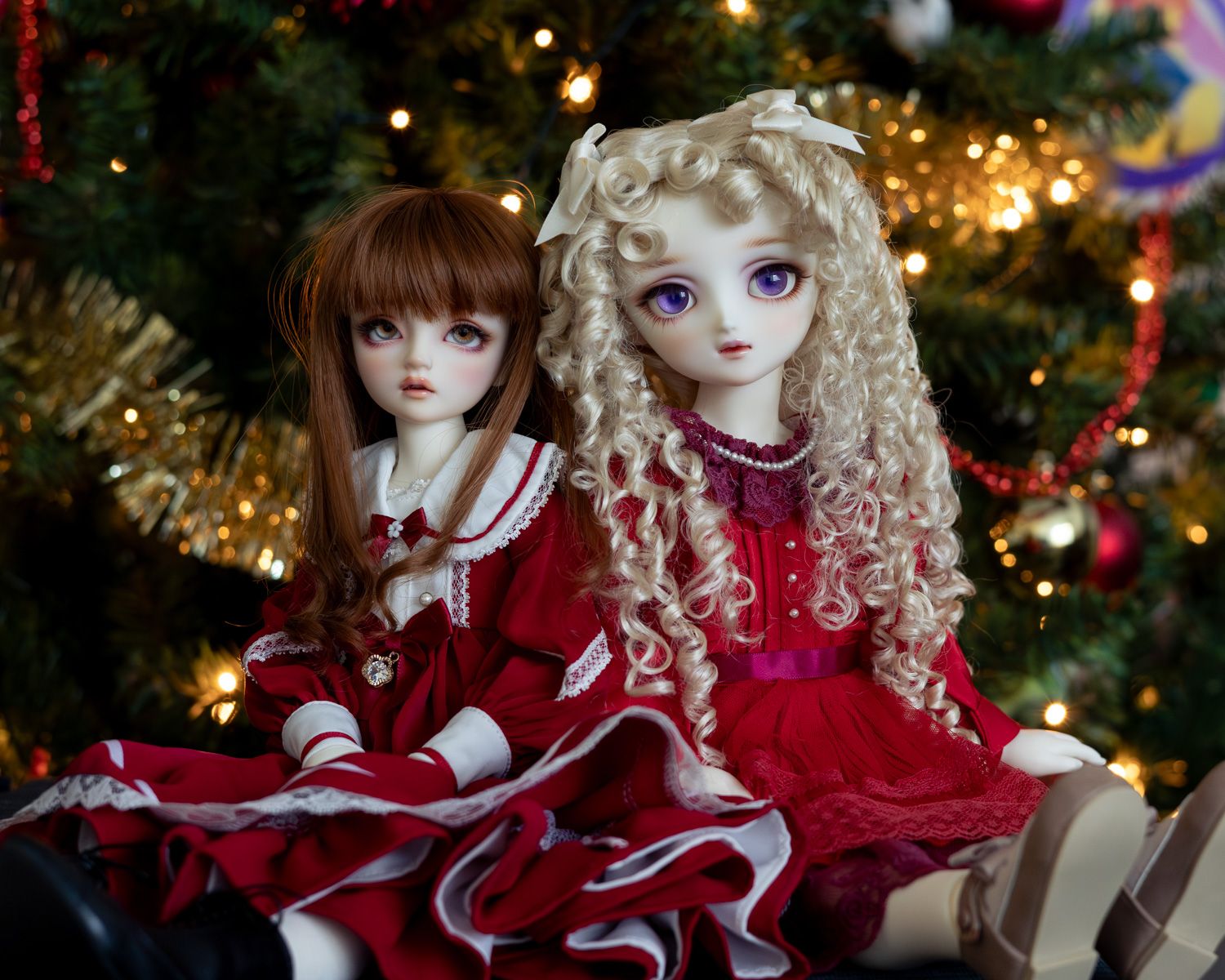 Two female ball-jointed dolls sit in front of a Christmas tree. They are both wearing red dresses. The doll on the left is smaller, with long auburn hair and a wistful expression. The doll on the right is larger, with big purple eyes and long curly blonde hair.