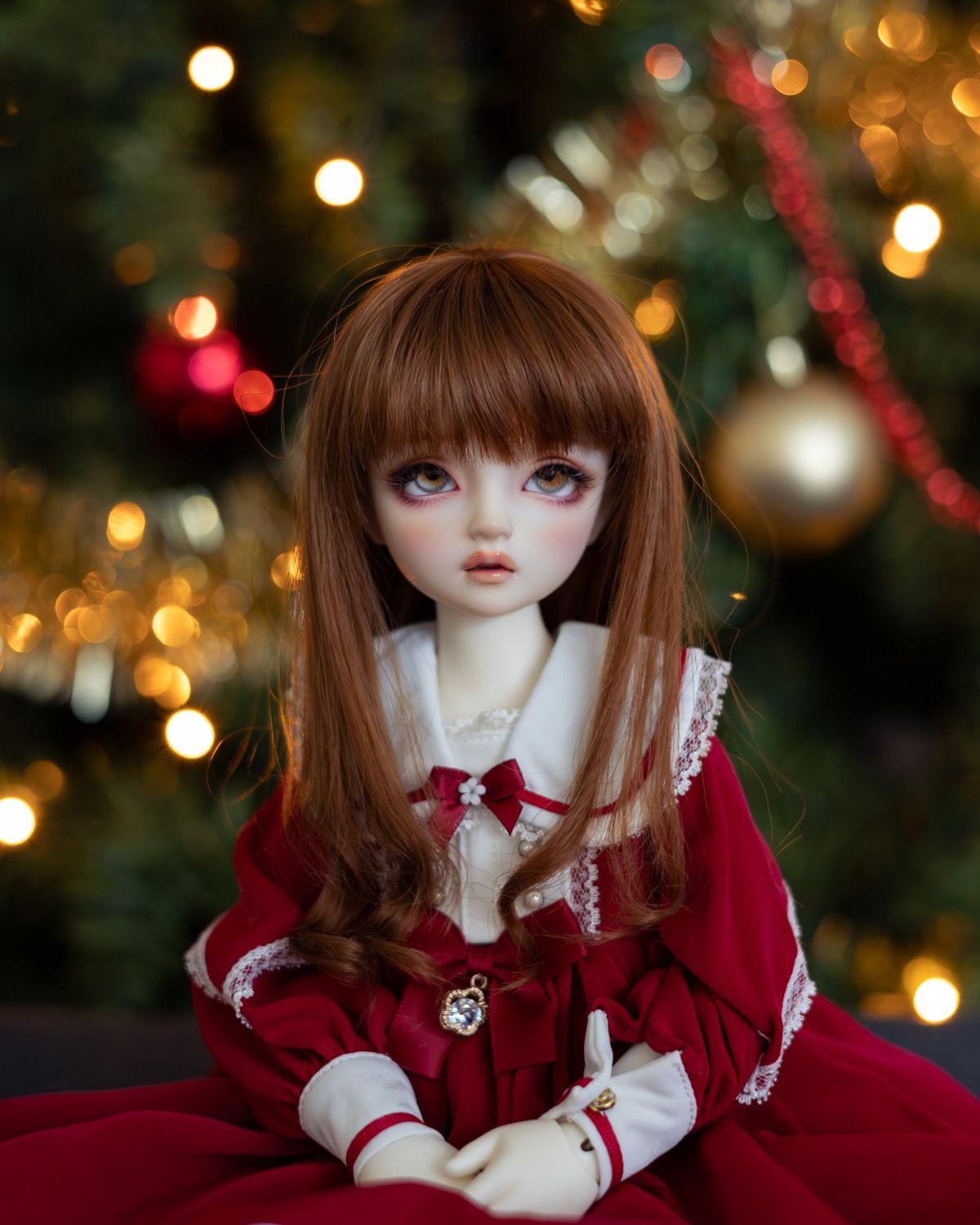 Half-length portrait of a female ball-jointed doll with long auburn hair, wearing a red and white dress with a large white sailor-like collar. She is sitting with her hands in her lap in front of an out-of-focus Christmas tree.