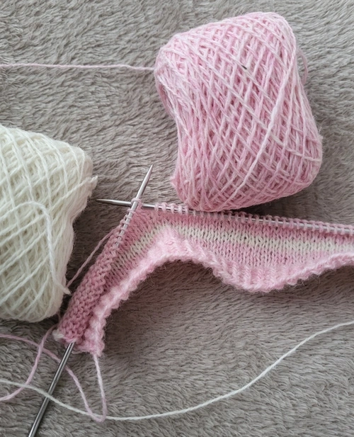 A piece of knitting in progress on knitting needles, using a ball of pink yarn and a ball of cream yarn.