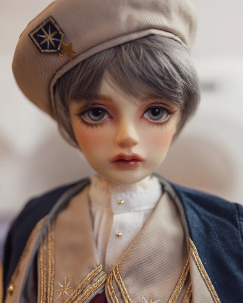 Portrait photo of a boyish-looking male resin ball-jointed doll, who is looking into the camera. He has thick eyelashes, short grey hair, grey eyes. He's wearing a beige beret with a star emblem, a white skirt with lace at the collar, and a dark blue and beige jacket with star emblems and gold trim.