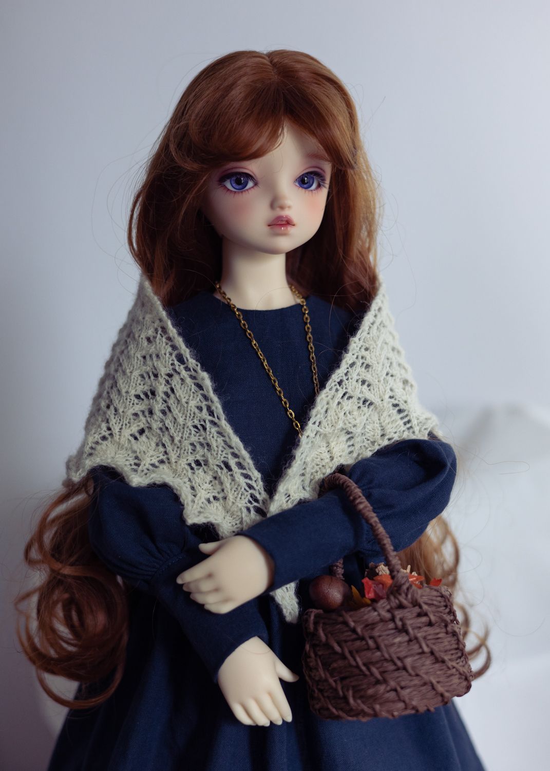 3/4 photo of a female ball-jointed doll wearing a navy dress with long sleeves, a cream lace shawl, a long pendant, and carrying a woven basket tucked over one arm. She has long wavy auburn hair, dark blue eyes, and stands against a light background.