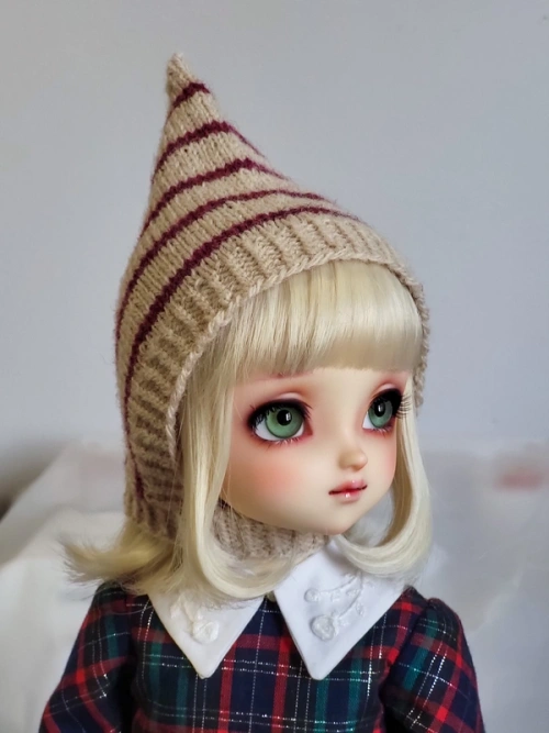 Three-quarter portrait photo of a female ball-jointed doll with big green eyes and a blond bob with blunt-cut fringe, wearing a knitted pointed pixie hat that covers her ears. The hat is oat-coloured and striped with burgundy.