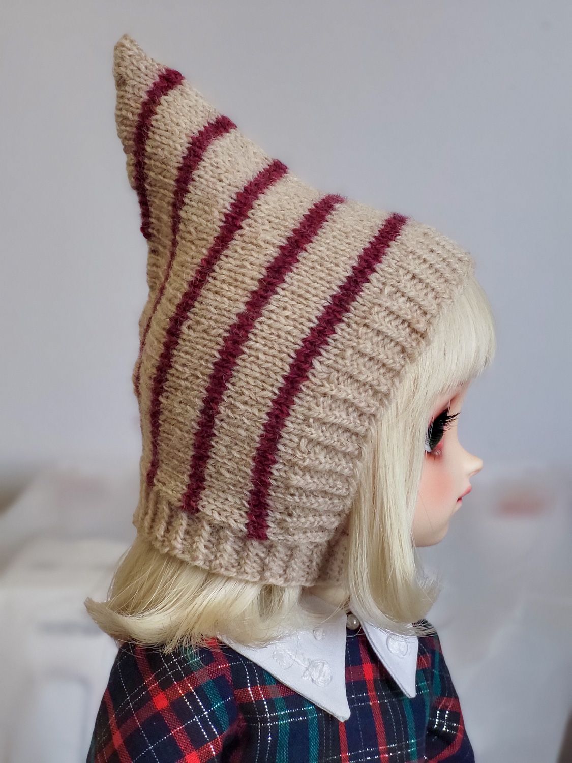 Side-profile photo of a female ball-jointed doll with a blond bob with blunt-cut fringe, wearing a knitted pointed pixie hat that covers her ears. The hat is oat-coloured and striped with burgundy.