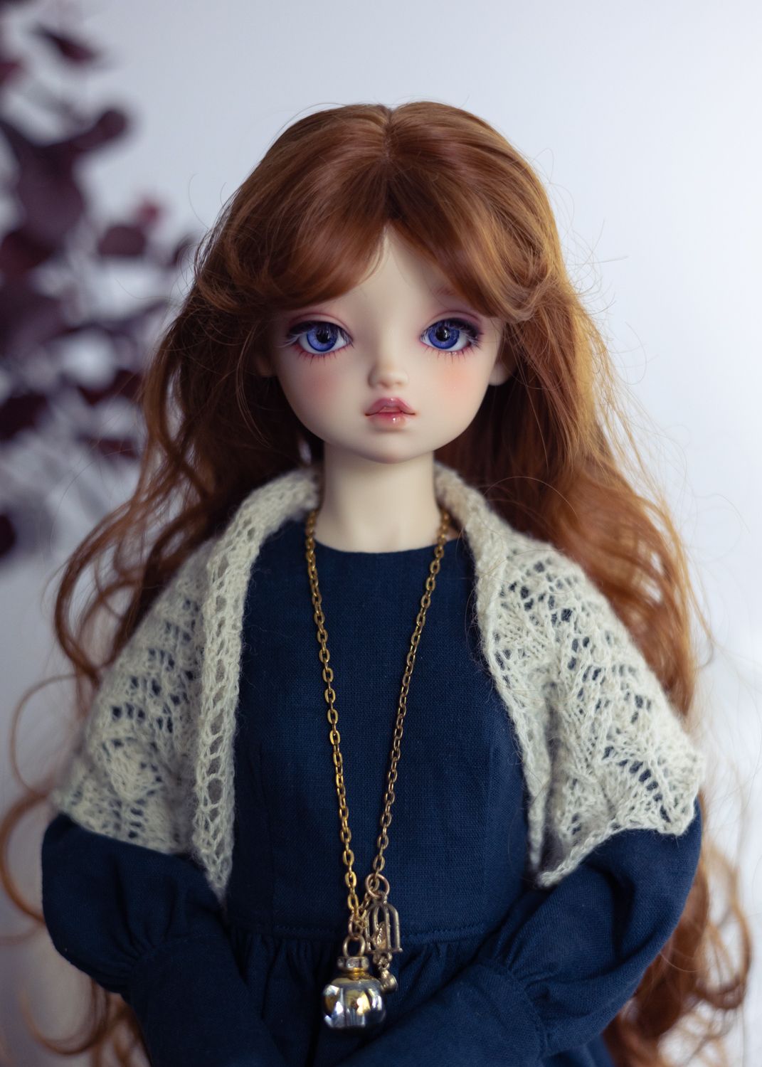 A portrait photo of a female ball-jointed doll wearing a navy dress with long sleeves, a cream lace shawl, and a long pendant. She is looking directly into the camera and has long wavy auburn hair, dark blue eyes, and stands against a light background.