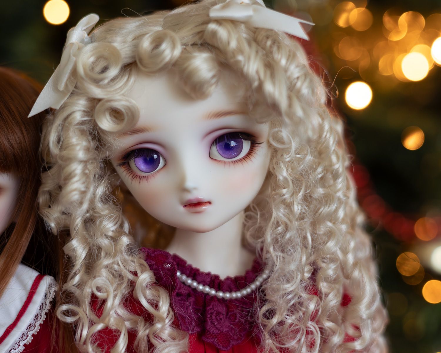 Portrait of a female ball-jointed doll with long blonde hair in ringlets and big sparkling purple eyes, wearing a red dress. She is in front of an out-of-focus Christmas tree.