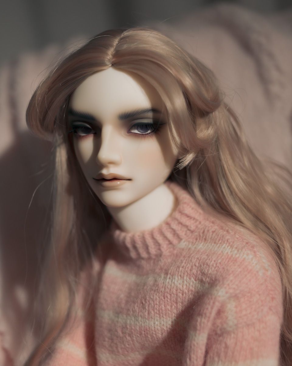 Portrait photo of a male ball-jointed doll with pale skin and long ash-pink hair. He wears a pink and white striped knitted sweater, and is looking into the camera.