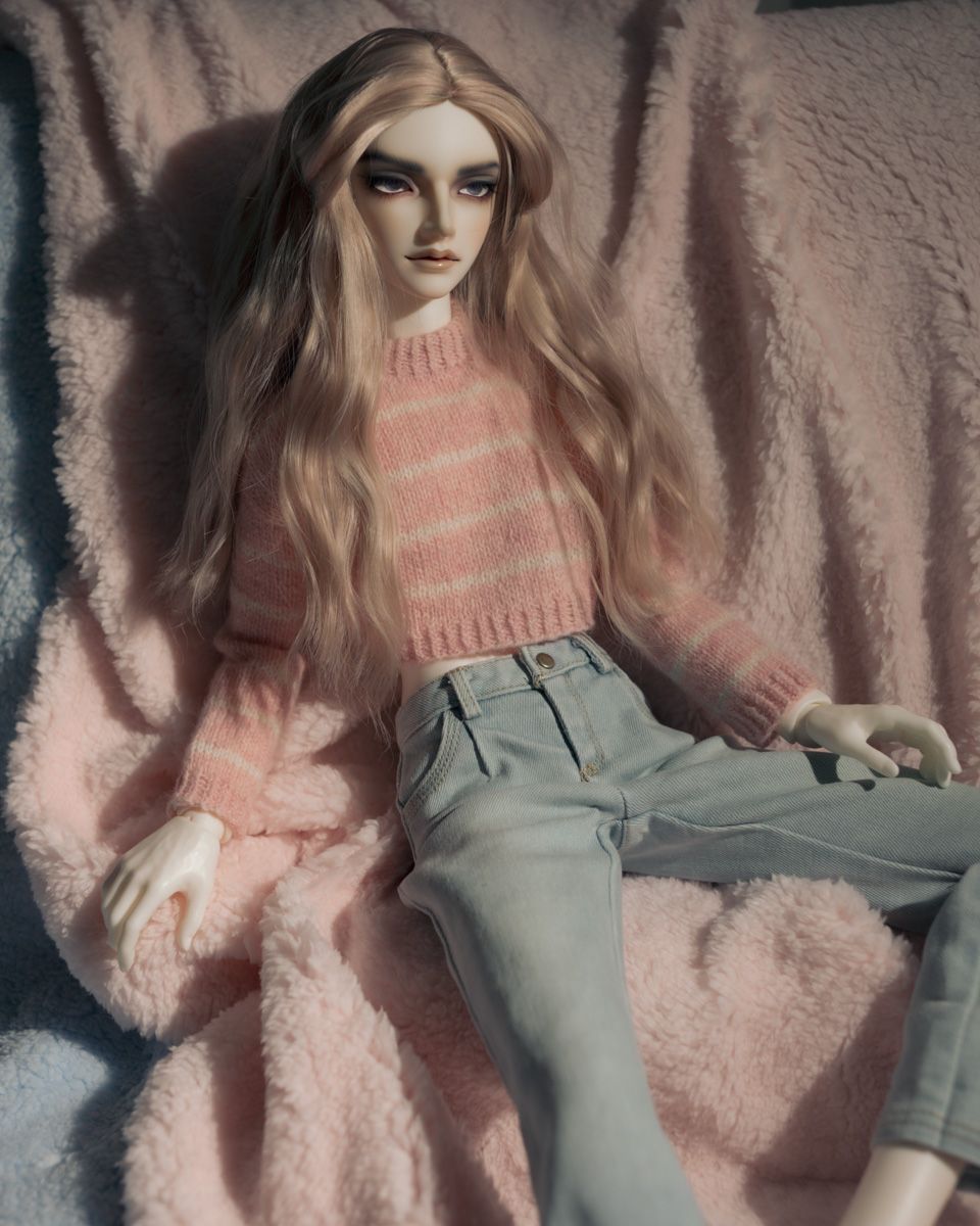 Male ball-jointed doll with pale skin and long ash-pink hair reclines on a pink and blue fur backdrop. He wears a pink and white striped knitted sweater and pale blue jeans, and is looking away from the camera.
