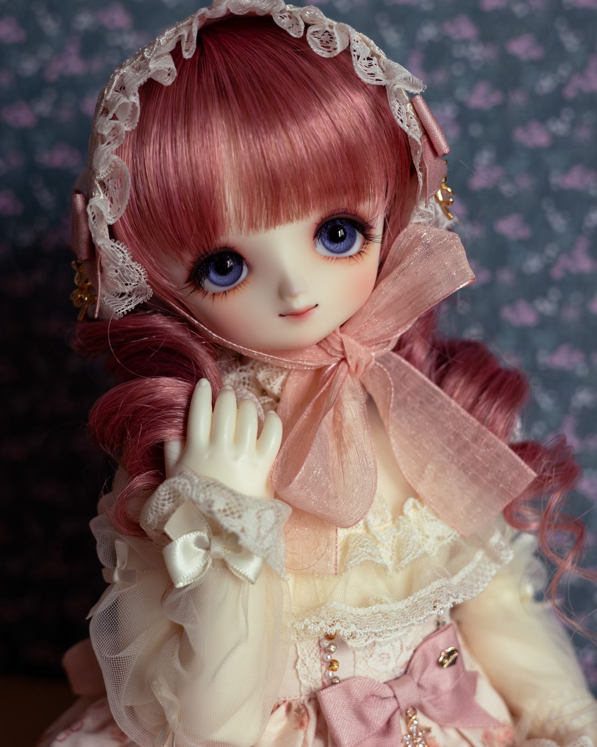 Half-body photo of a female ball-jointed doll with big blue eyes, a pink and white frilly outfit, and a pink wig with curls. Her hand is touching one of the curls.