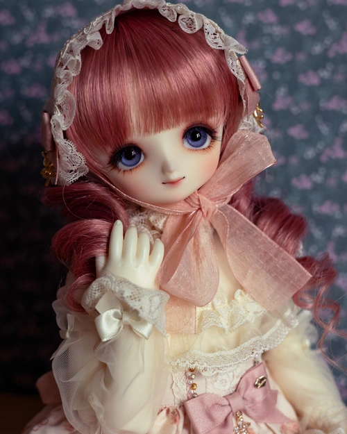 Half-body photo of a female ball-jointed doll with big blue eyes, a pink and white frilly outfit, and a pink wig with curls. Her hand is touching one of the curls.