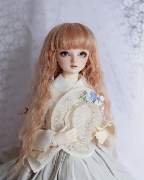 Three-quarter portrait of a female ball-jointed doll in an old-fashioned looking blouse and skirt, holding a mesh hat trimmed with small fake flowers. She has long wavy peach-orange hair with a thick fringe, and a slight smile.