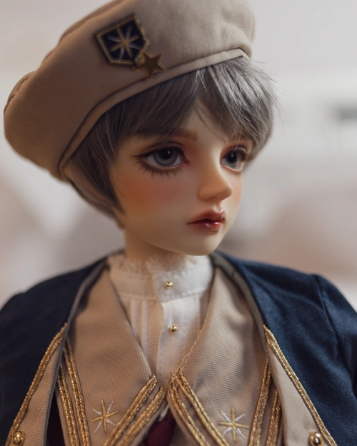 Three-quarter portrait photo of a boyish-looking male resin ball-jointed doll. He has thick eyelashes, short grey hair, grey eyes. He's wearing a beige beret with a star emblem, a white skirt with lace at the collar, and a dark blue and beige jacket with star emblems and gold trim.