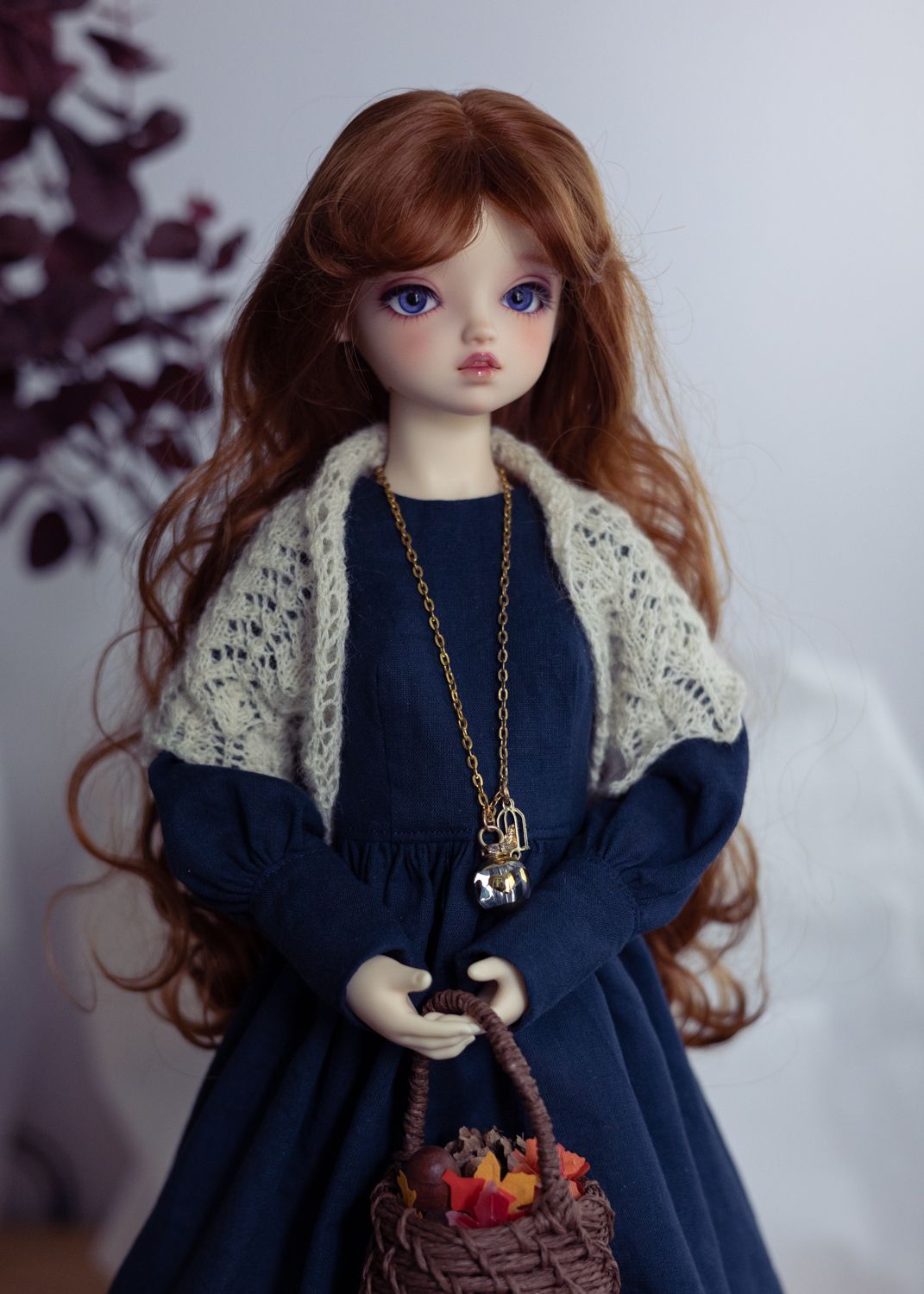 3/4 photo of a female ball-jointed doll wearing a navy dress with long sleeves, a cream lace shawl, a long pendant, and carrying a woven basket. She has long wavy auburn hair, dark blue eyes, and stands against a light background.