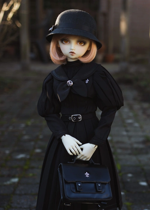 Photo of a ball-jointed resin doll. She is outside in a garden with grey tiles behind her, wearing a black old-fashioned school uniform and a cloche hat. She holds a black school bag in front of her. Her resin is pale white, her eyes are orange and her bobbed hair is peach.