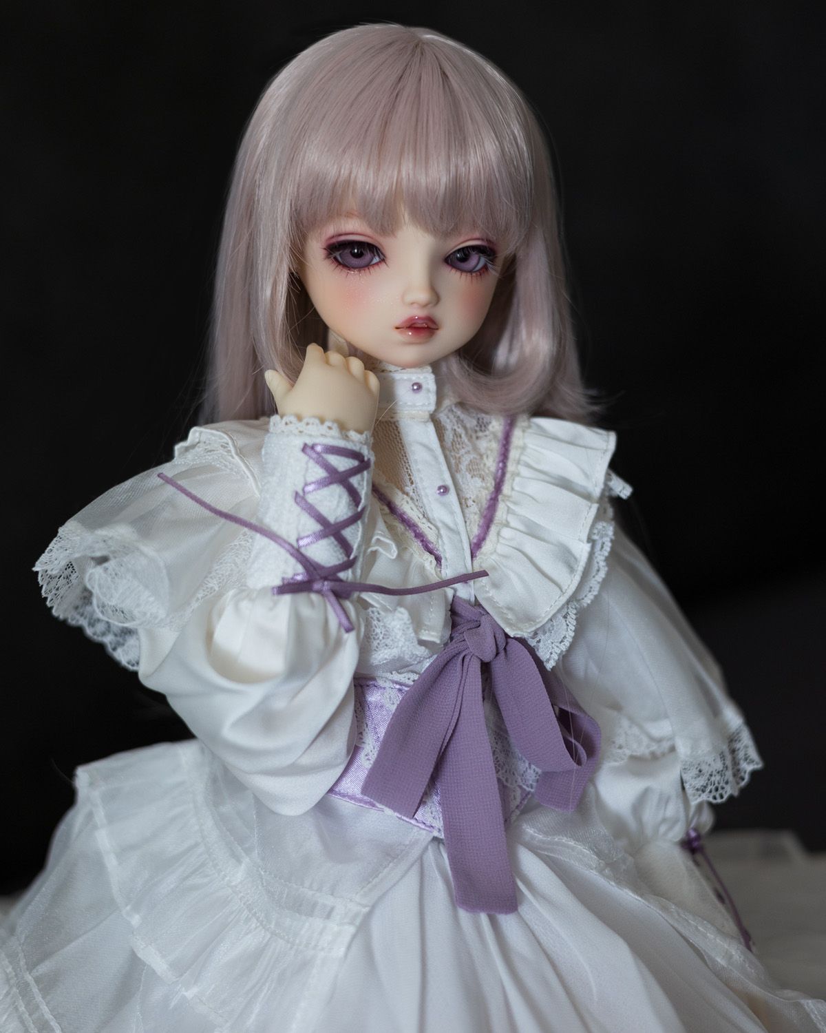 Female ball-jointed doll with a superior expresion, a light desaturated pink wig, and a frilled white dress with purple ribbon accents.